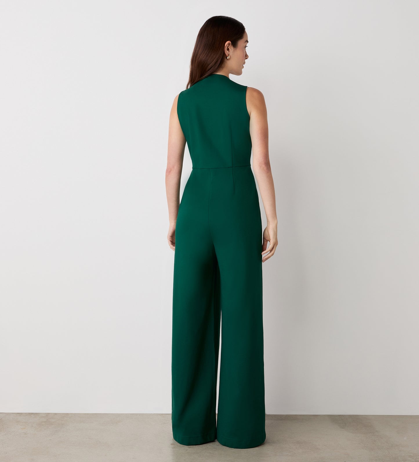 A woman stands with her back to the camera, wearing the Effie Bottle Green Ponte Jersey Jumpsuit—featuring wide legs and a sleeveless v-neck design—against a plain light-colored wall and concrete floor.