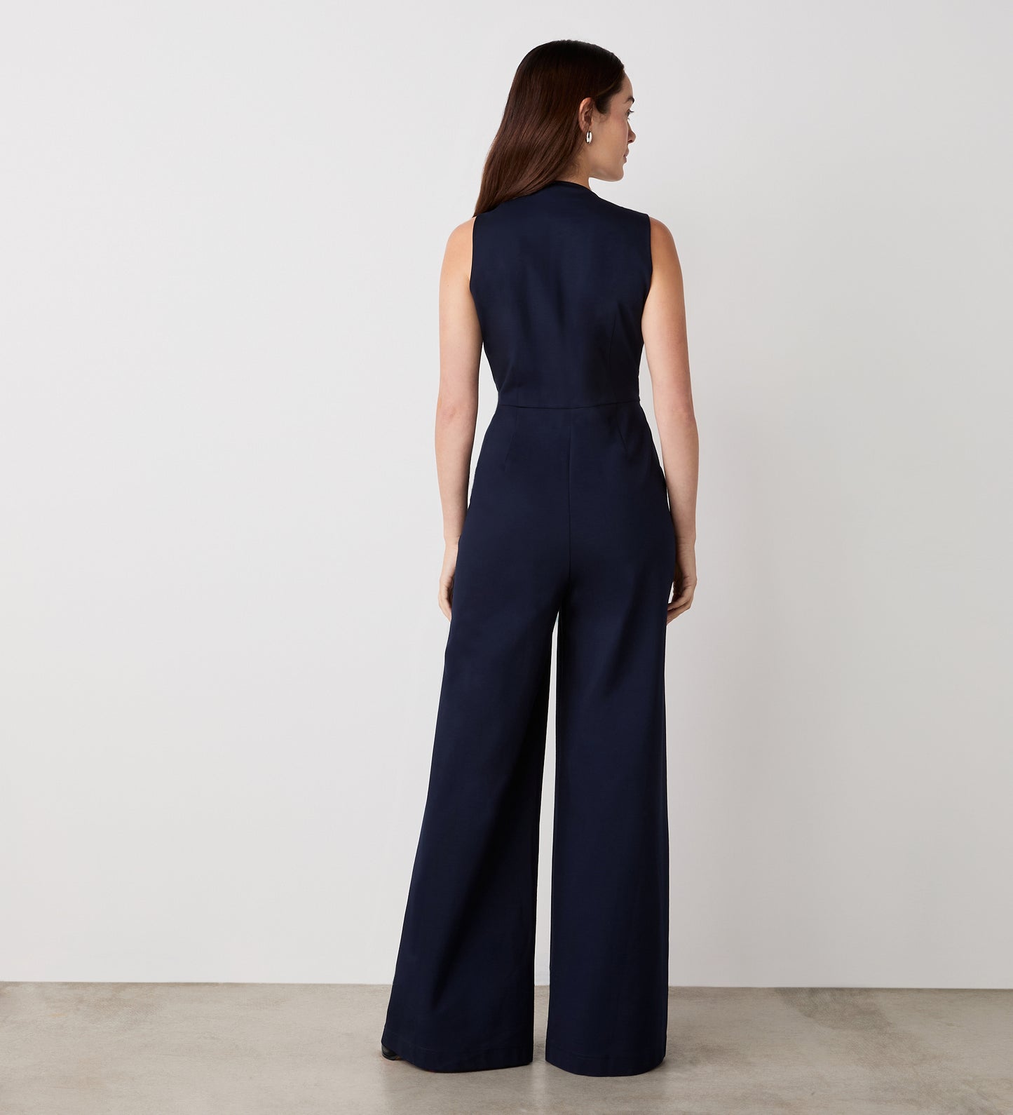 A woman with long brown hair faces away, wearing the Effie Navy Ponte Jersey Jumpsuit. The sleeveless, wide-leg outfit is styled indoors against a plain white wall and light-colored floor.