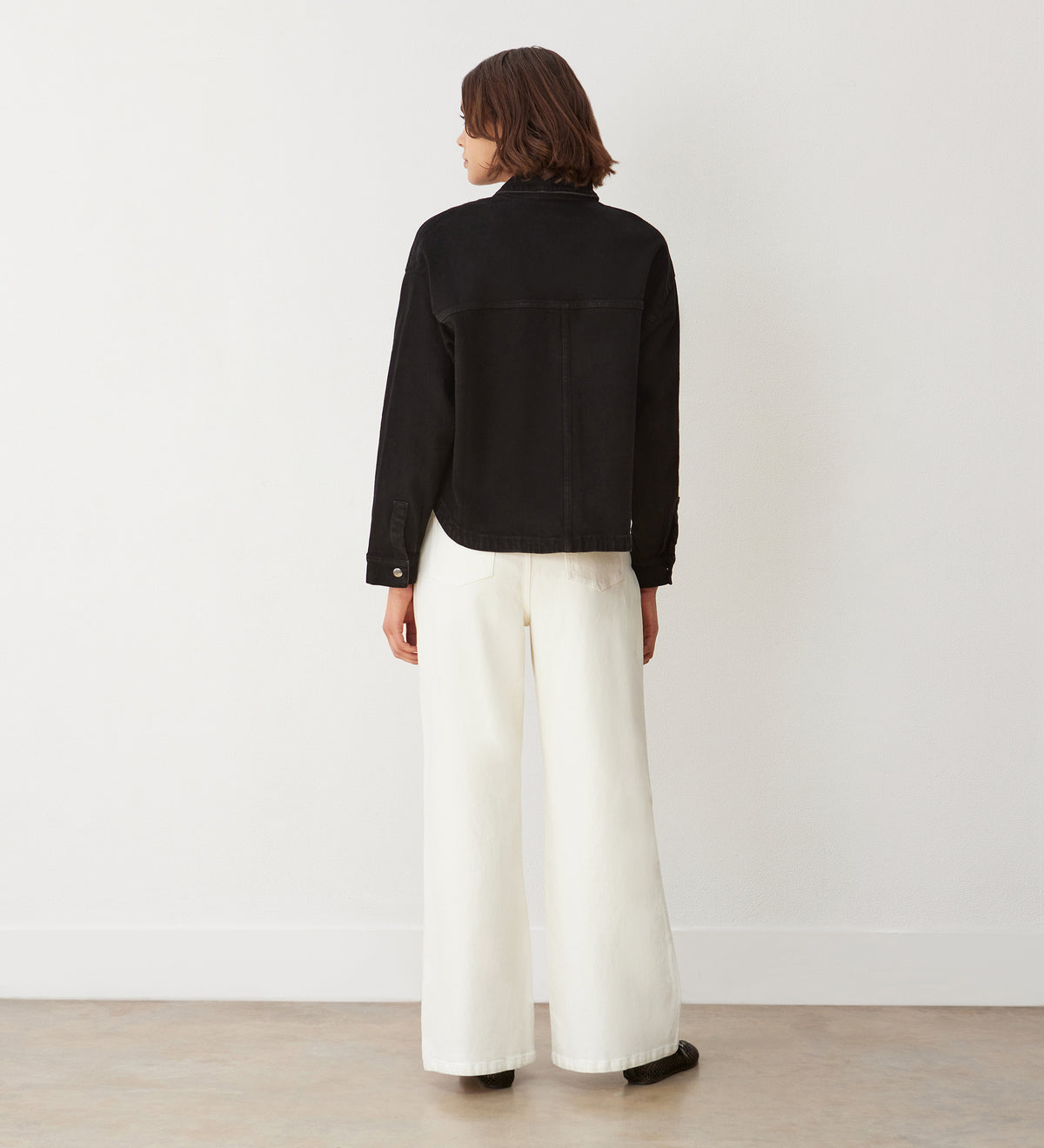 A person with short brown hair stands facing a white wall, wearing the Elidih Black Denim Jacket with front patch pockets, wide-leg white pants, and black shoes—an essential indoor wardrobe look on a light-colored floor.