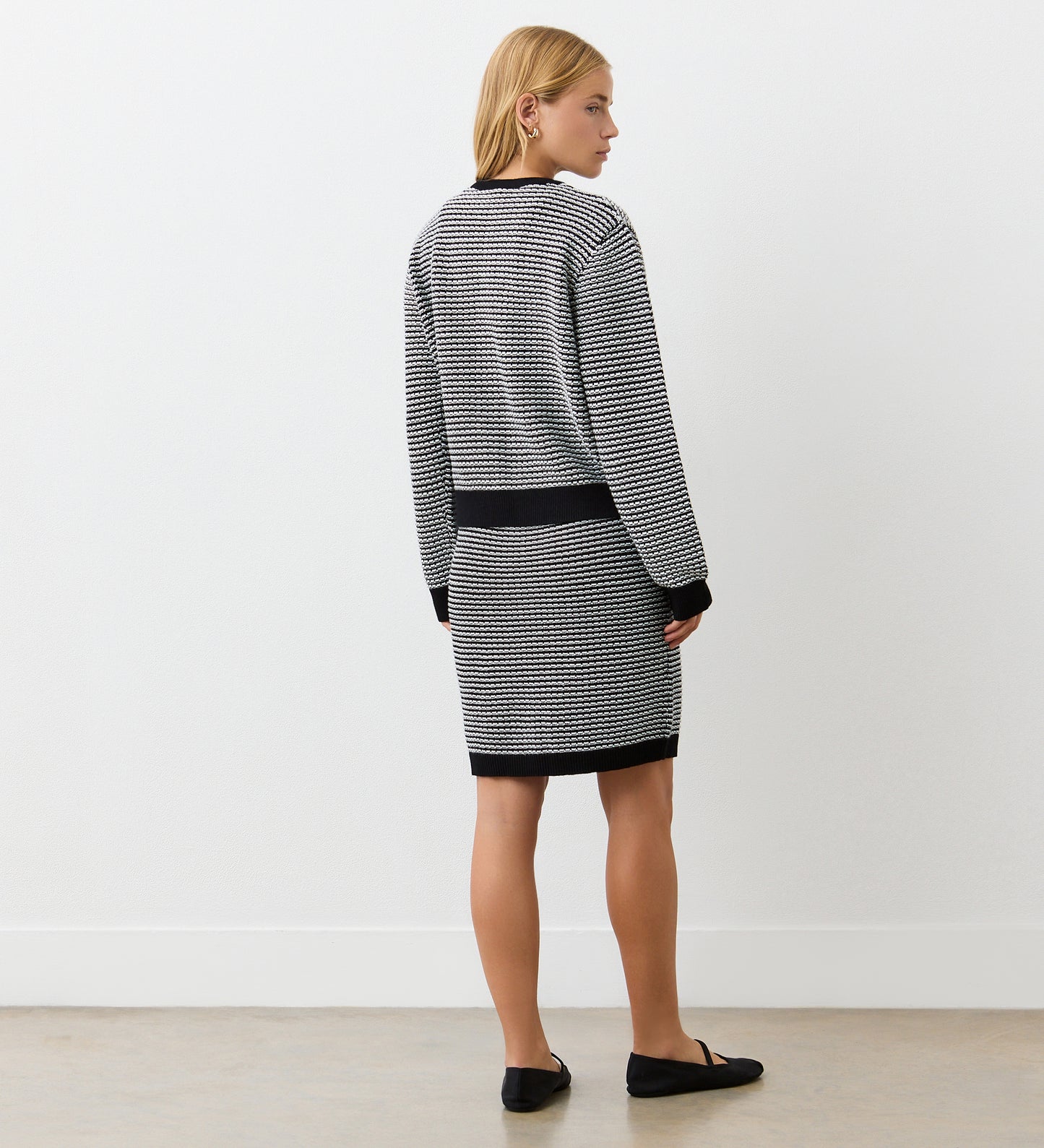 A woman with blonde hair, indoors against a white wall on a light wood floor, wears the Emery Black Knitted Cardigan with gold-rimmed buttons and a matching striped skirt trimmed in black, paired with black flats.