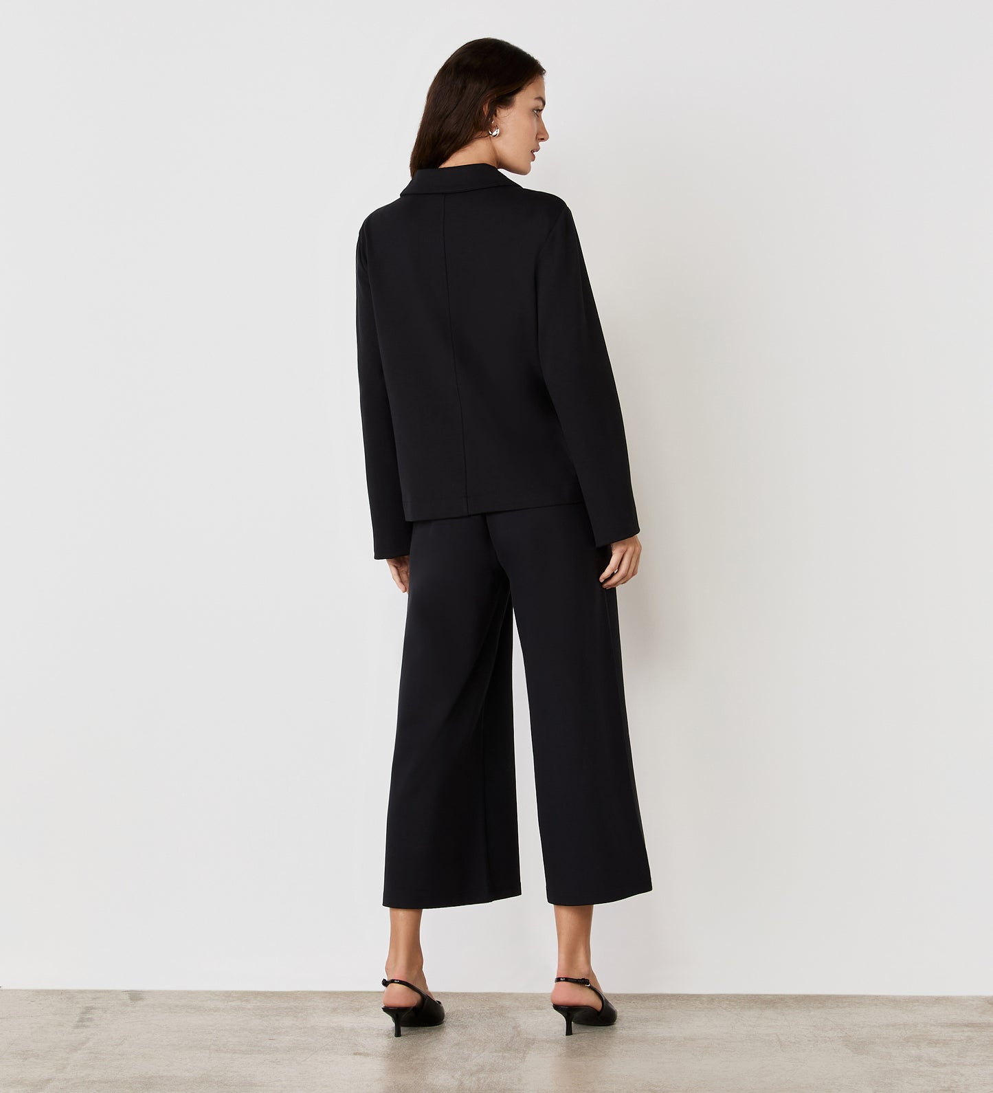 A woman faces away from the camera wearing the Enola Black Ponte Jersey Jacket with wide-leg cropped ponte jersey pants and black heeled sandals, set against a plain white background.