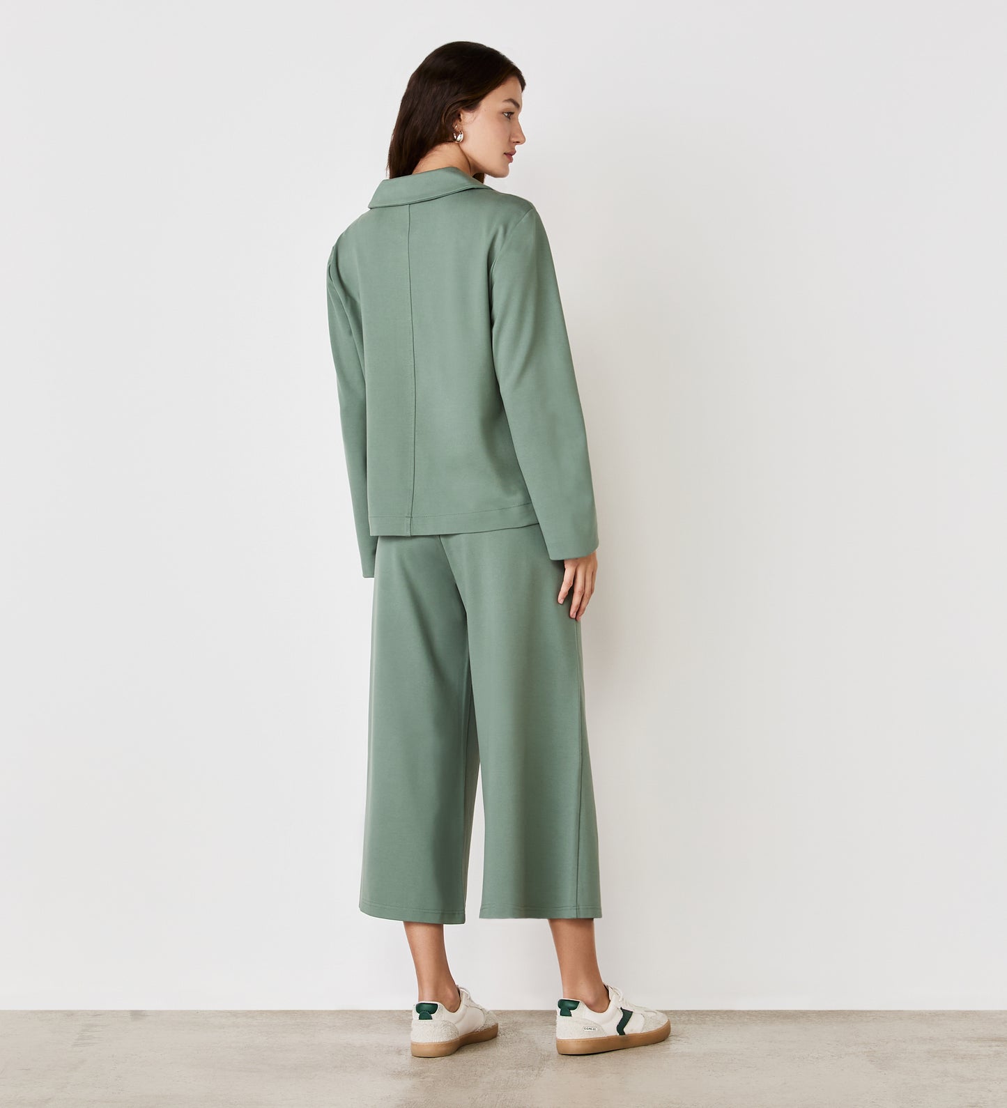 A woman faces away from the camera in the Enola Light Khaki Ponte Jersey Jacket with matching wide-leg cropped pants and white sneakers, standing against a plain white wall on a light-colored floor.