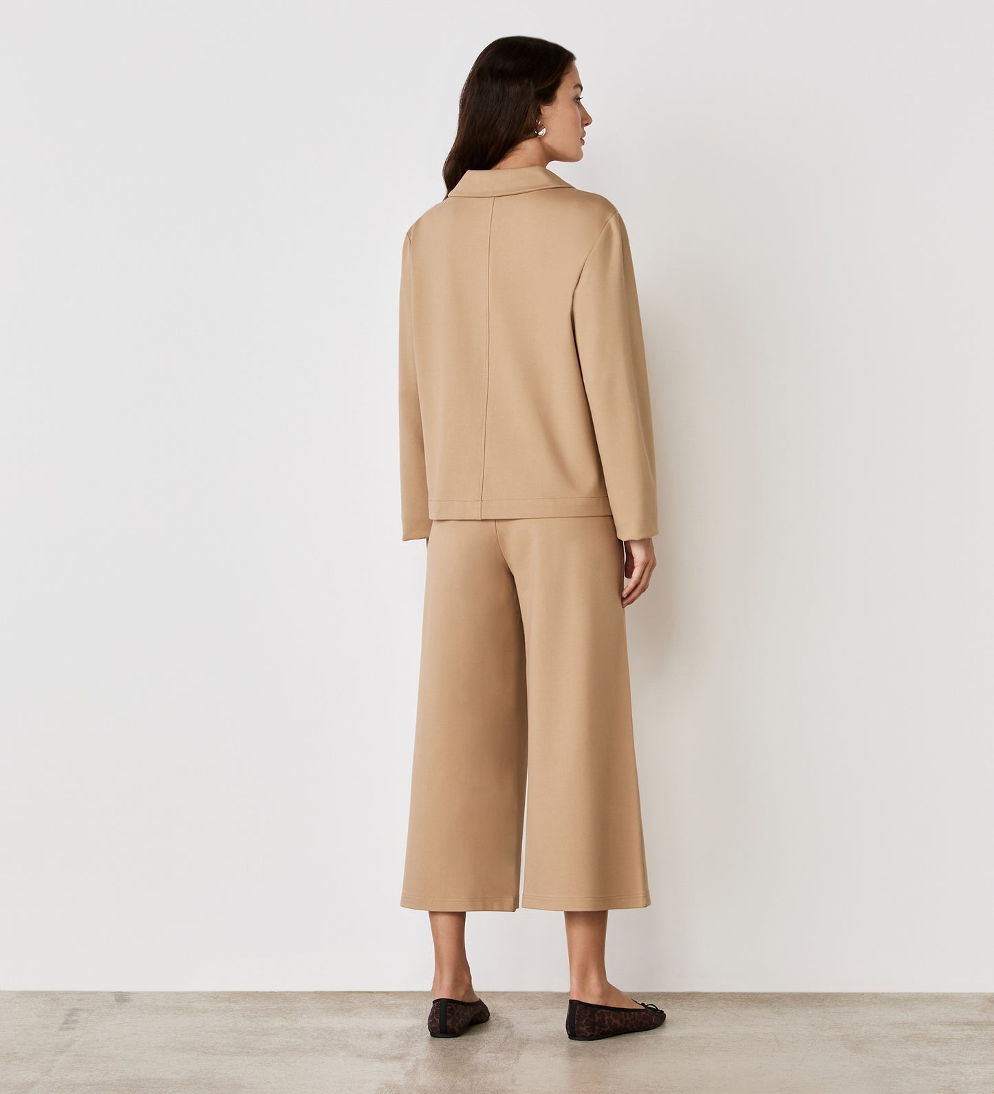A woman stands indoors on a concrete floor, facing away from the camera, wearing the Enola Beige Ponte Jersey Jacket with matching wide-leg cropped pants against neutral-colored walls.