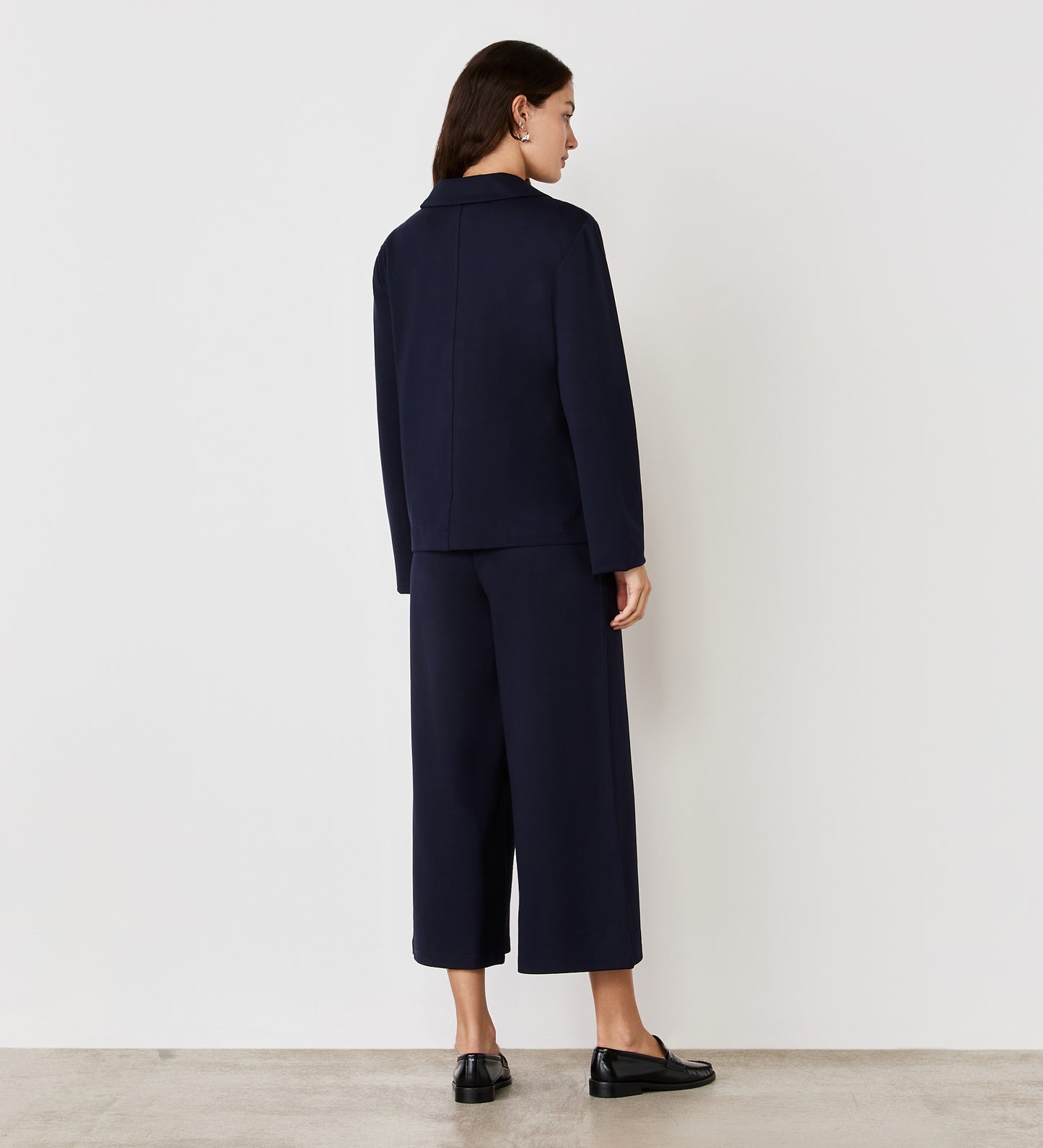 A woman faces away from the camera, wearing the Enola Navy Ponte Jersey Jacket with matching wide-leg trousers and black loafers, set against a plain white wall and concrete floor.