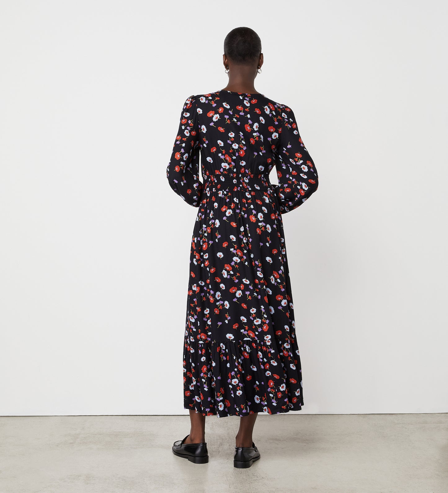 A person faces a white wall wearing the Erina Black Floral Midi Dress, an ankle-length, long-sleeved dress with red and white floral prints—a striking choice for any floral collection—paired with classic black shoes.