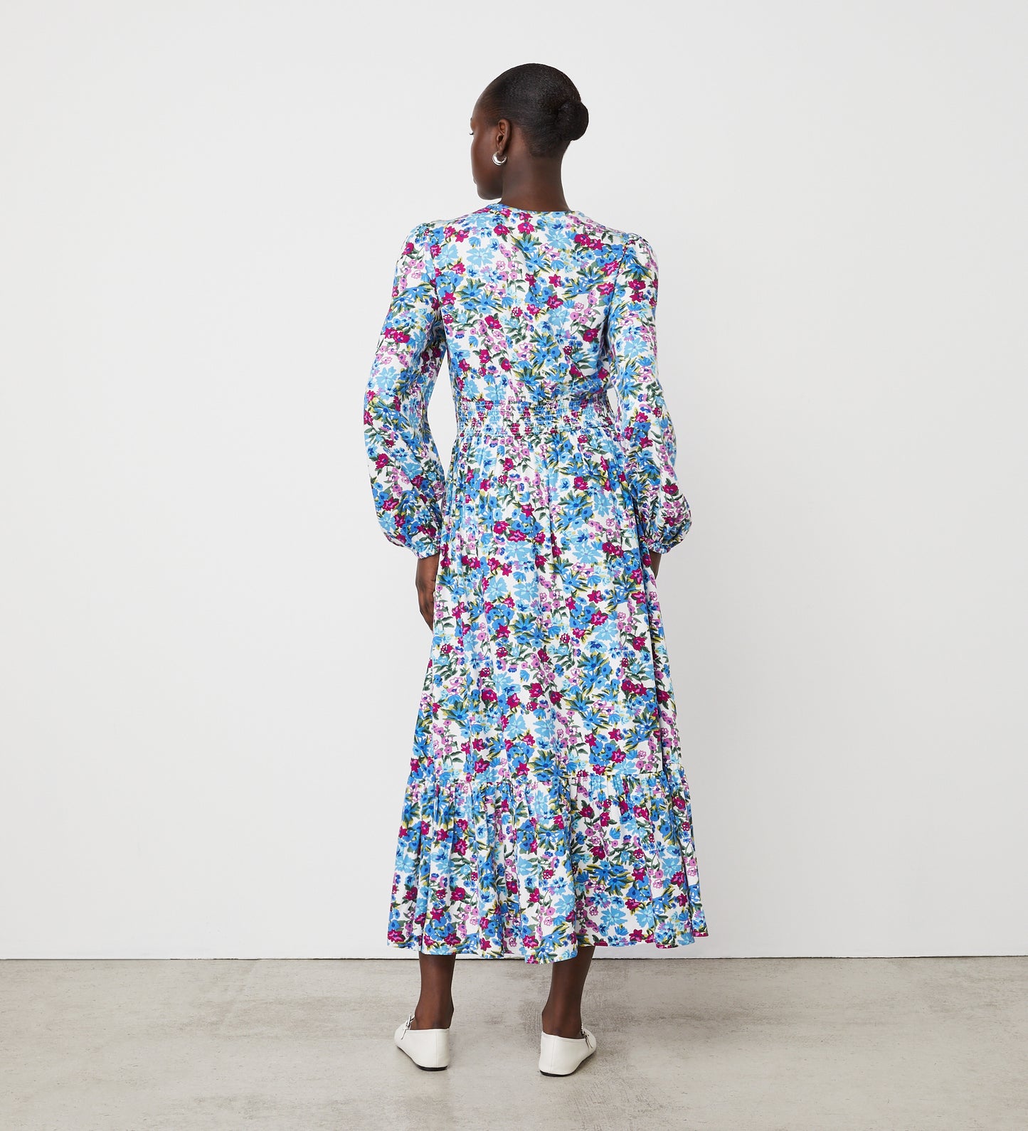 A woman with her hair in a bun stands facing away on a plain white background, wearing the Erina Blue Floral Midi Dress featuring vibrant blue, purple, and pink florals with a shirred empire waist, paired with white shoes.