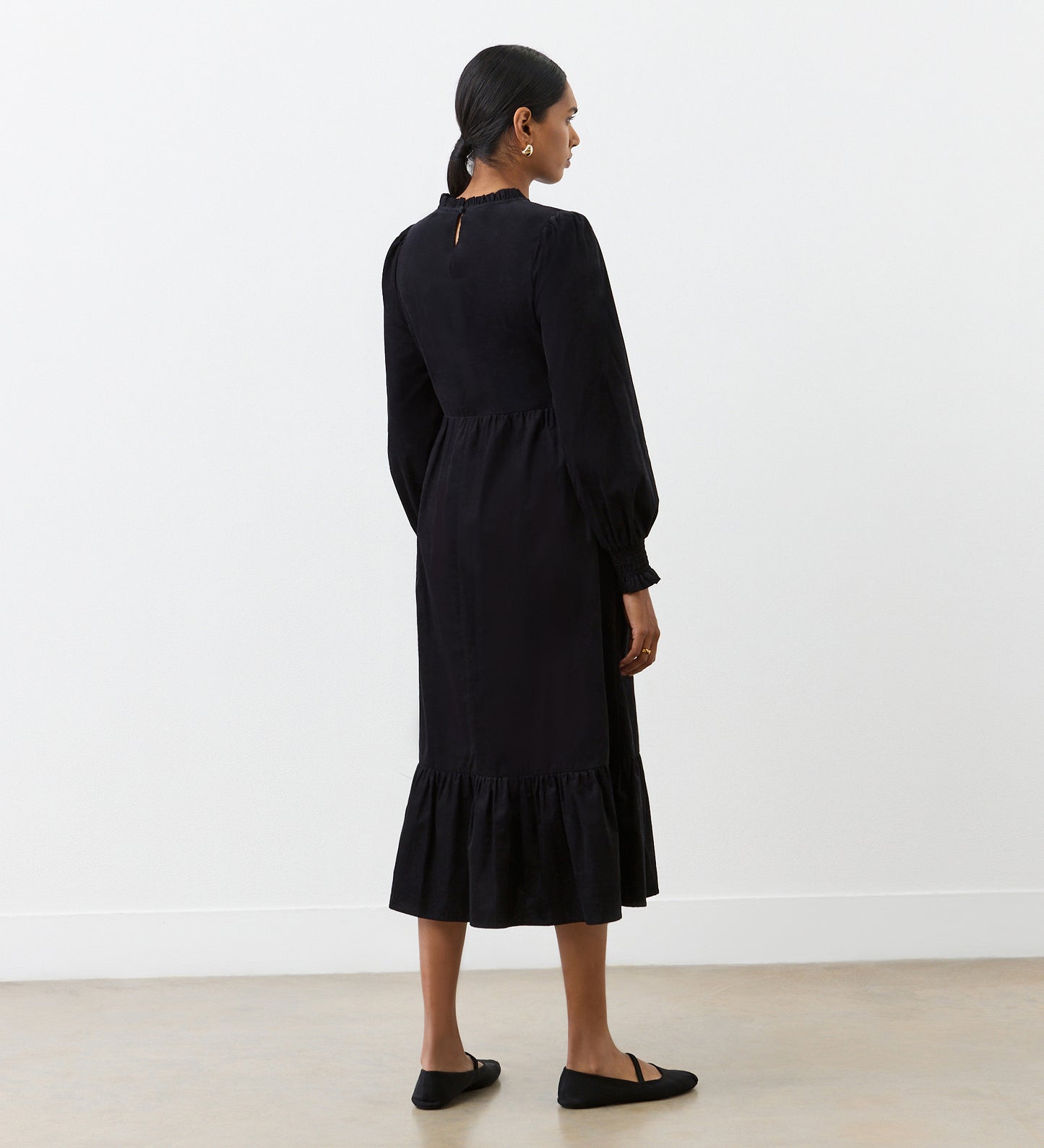 A woman with a low ponytail stands against a white wall wearing the Esme Black Corduroy Midi Dress, featuring a ruffled, tiered hemline, paired with simple black flats.