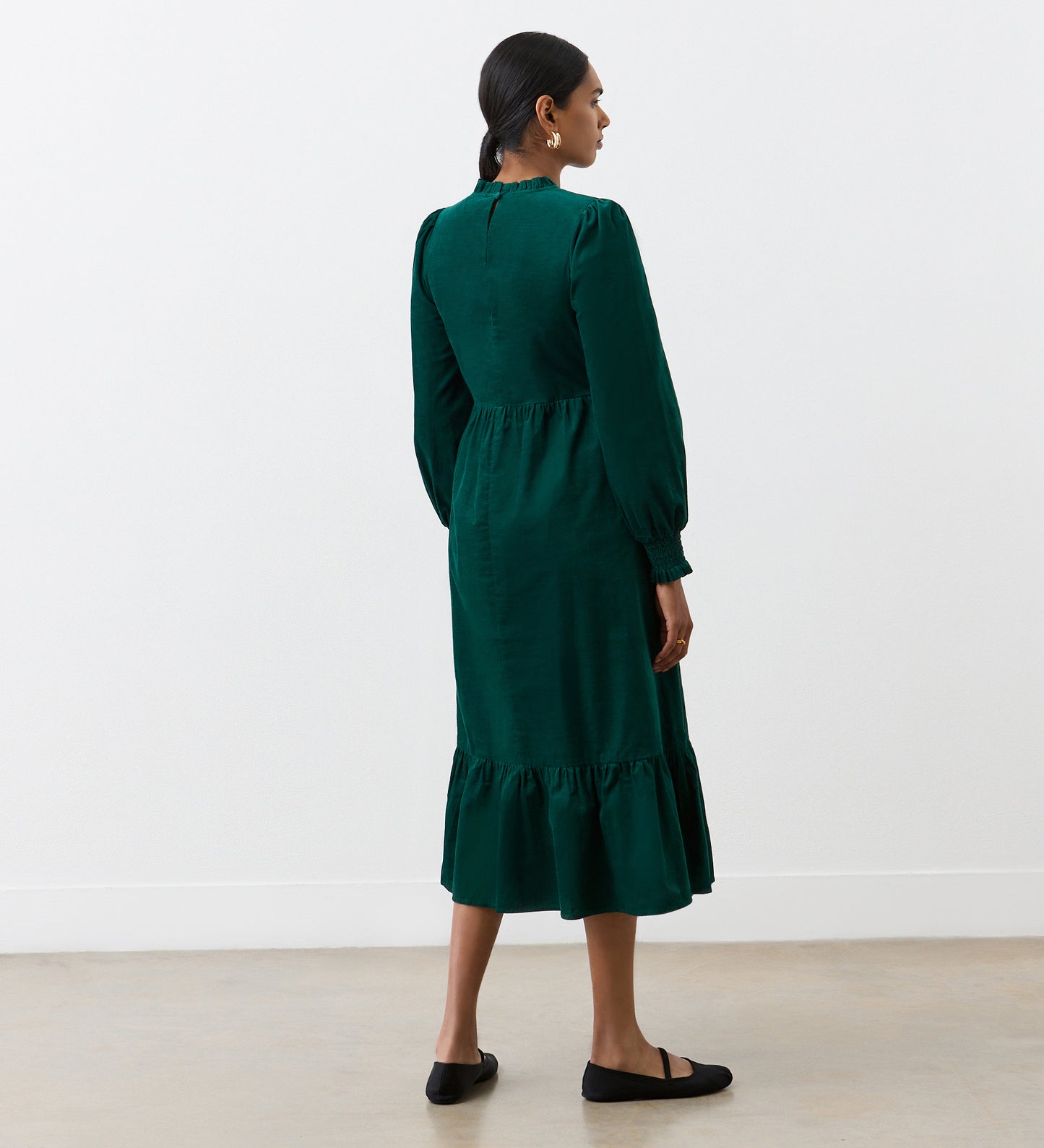 A woman faces away, wearing the Esme Bottle Green Corduroy Midi Dress with long sleeves and a ruffled tiered hem, styled with black flats. Her hair is tied back as she stands in a minimalist room with light walls and a bare floor.