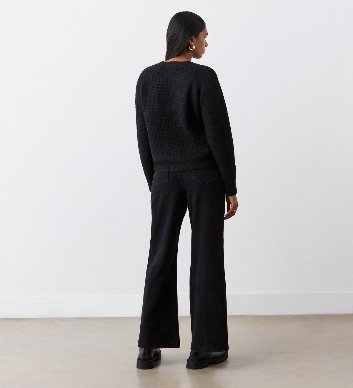 A person stands facing away from the camera, wearing the Etta Black Cardigan with long raglan sleeves, black wide-leg pants, and platform shoes in a minimalist room with white walls and a light floor.