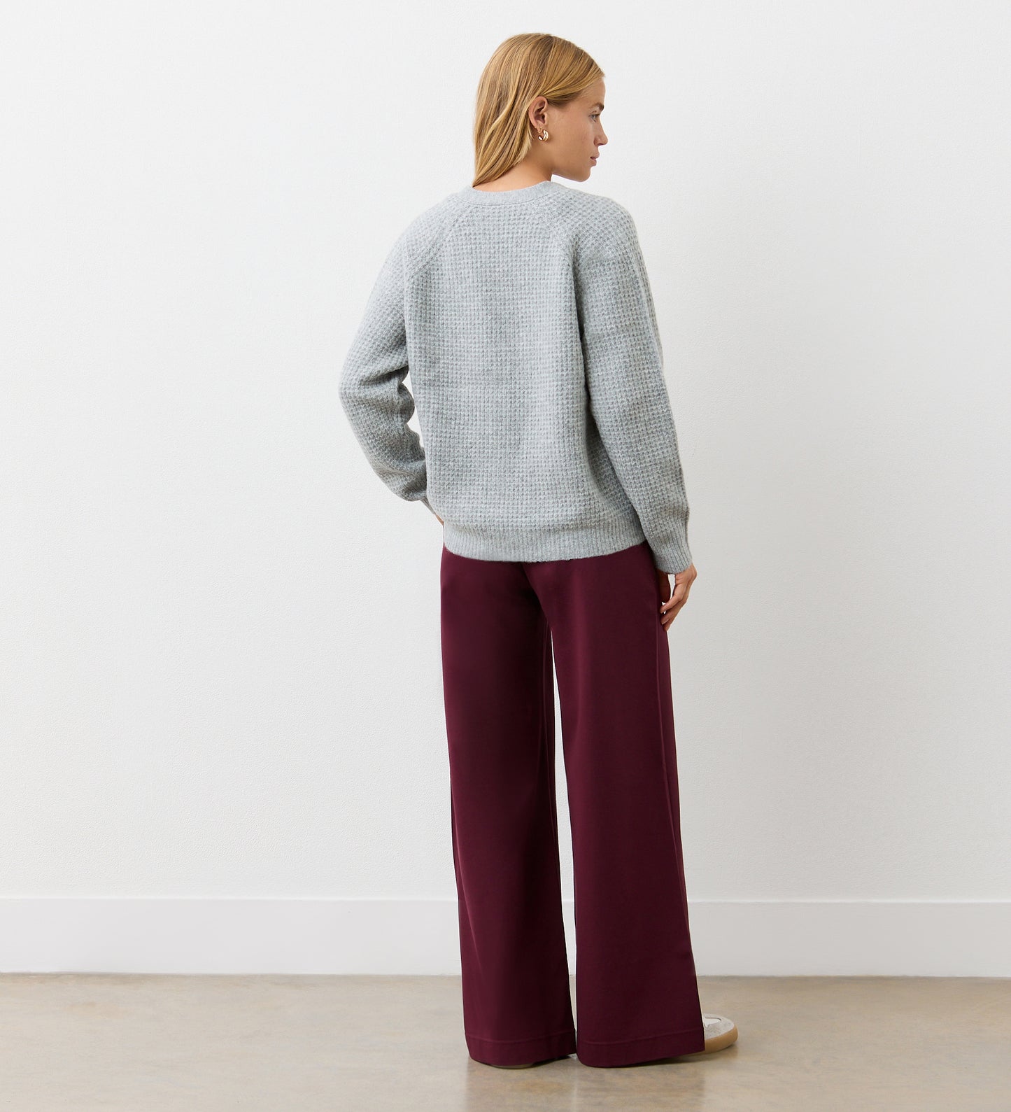 A woman faces away, wearing the Etta Grey Marle Cardigan with wide-legged burgundy pants. She stands by a plain white wall on a light floor, her right hand at her side and her left hand on her hip.