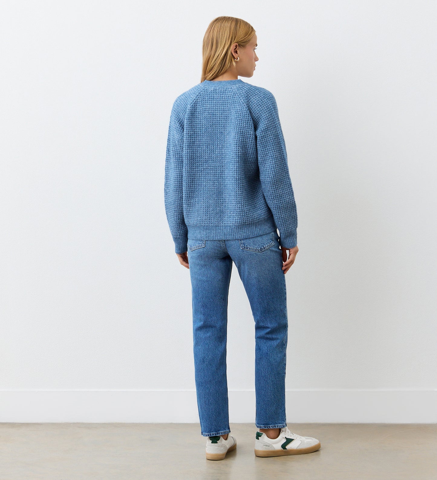 A person with blonde hair, wearing the Etta Blue Cardigan, blue jeans, and white sneakers stands facing away from the camera against a white wall and beige floor.