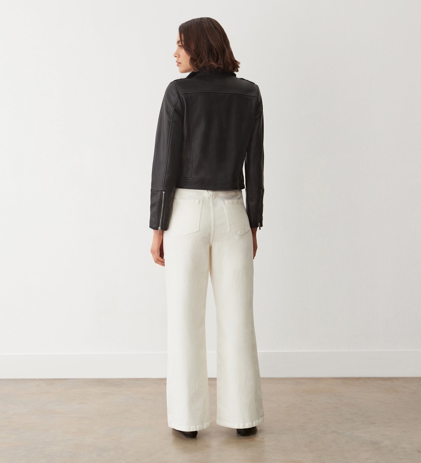 A person with short brown hair stands facing away, wearing the Faith Black Leather Biker Jacket with silver hardware and white wide-leg pants in a minimalist room with white walls and a concrete floor.