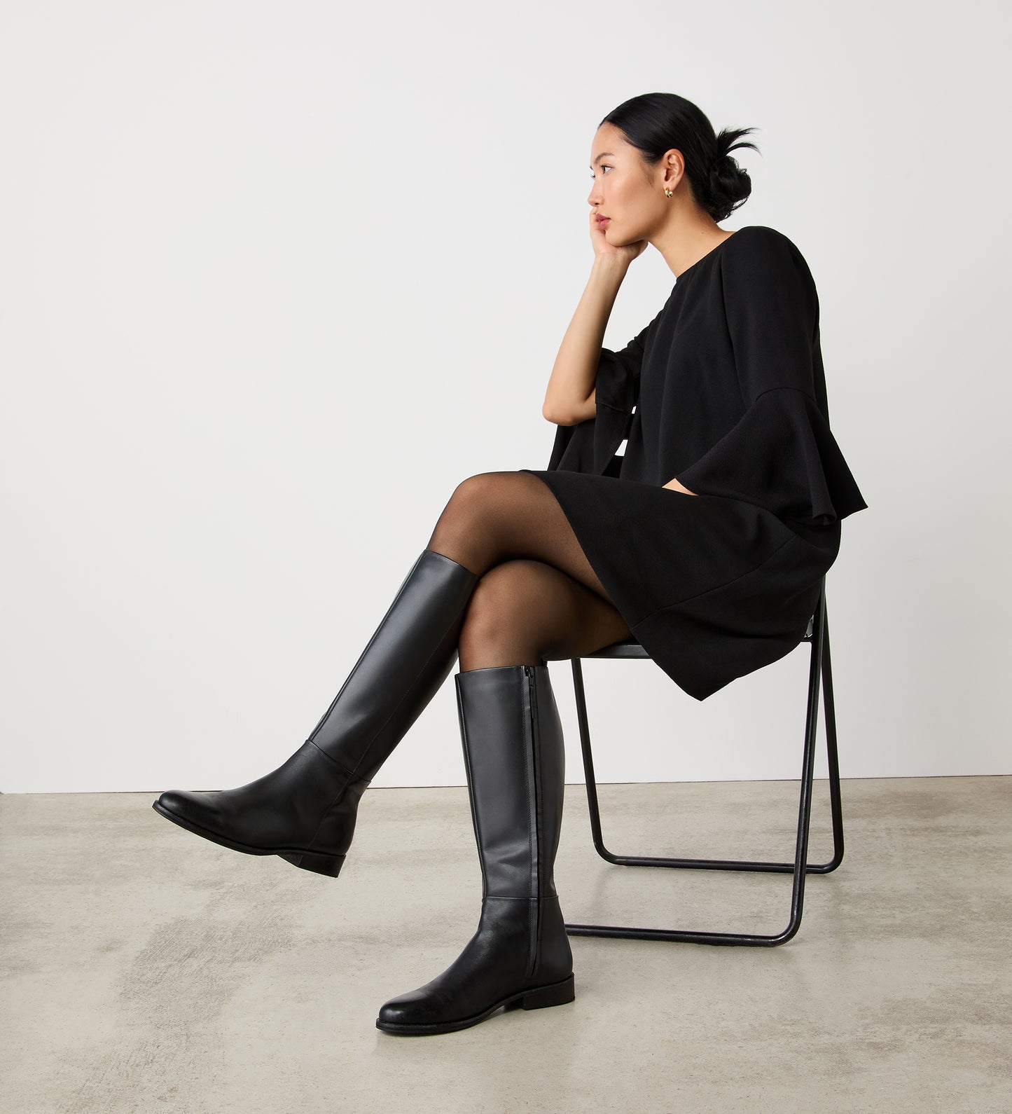 A woman wearing Frankie Black Knee High Leather Boots, a black dress, and sheer black tights sits sideways on a simple chair, gazing thoughtfully into the distance against a plain white wall and concrete floor.