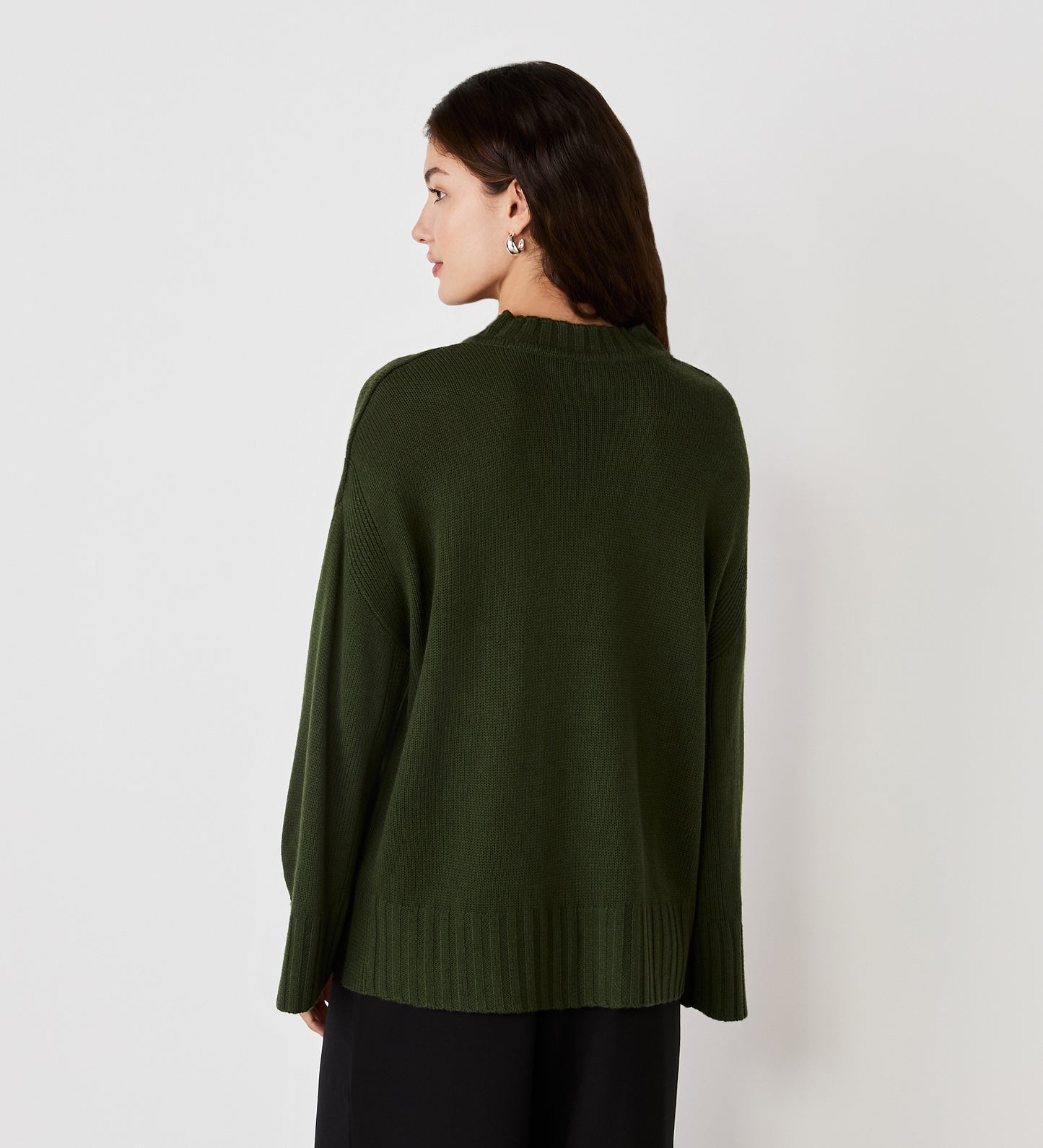 A woman with long dark hair faces away, wearing the Frankie Khaki Boxy Jumper with split cuffs and black pants against a plain white background.