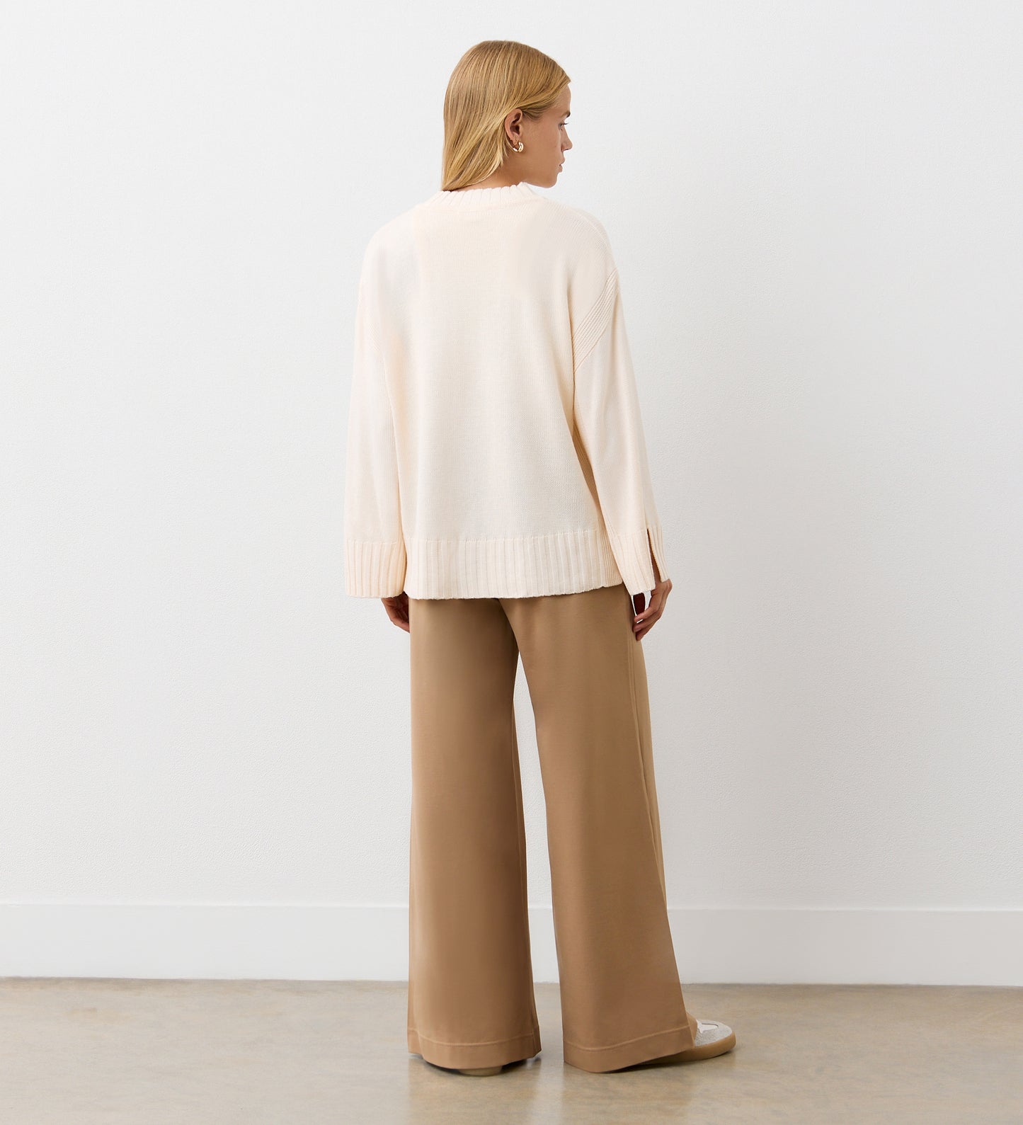 A woman with straight, shoulder-length blonde hair stands facing away in a minimalist room, wearing the Frankie Ivory Boxy Jumper and wide-leg tan trousers. The loose cream jumper features a round neck and deep ribbed hemline.