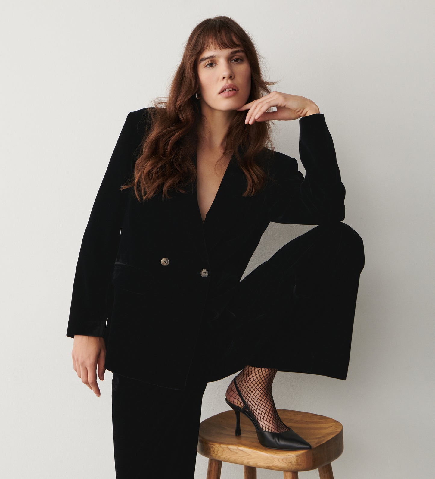 A woman with long brown hair wears the Maeve Black Velvet Blazer, styled with a structured fit and fishnet heels, as she sits on a wooden stool against a plain white background, one leg up and her elbow resting on her knee.