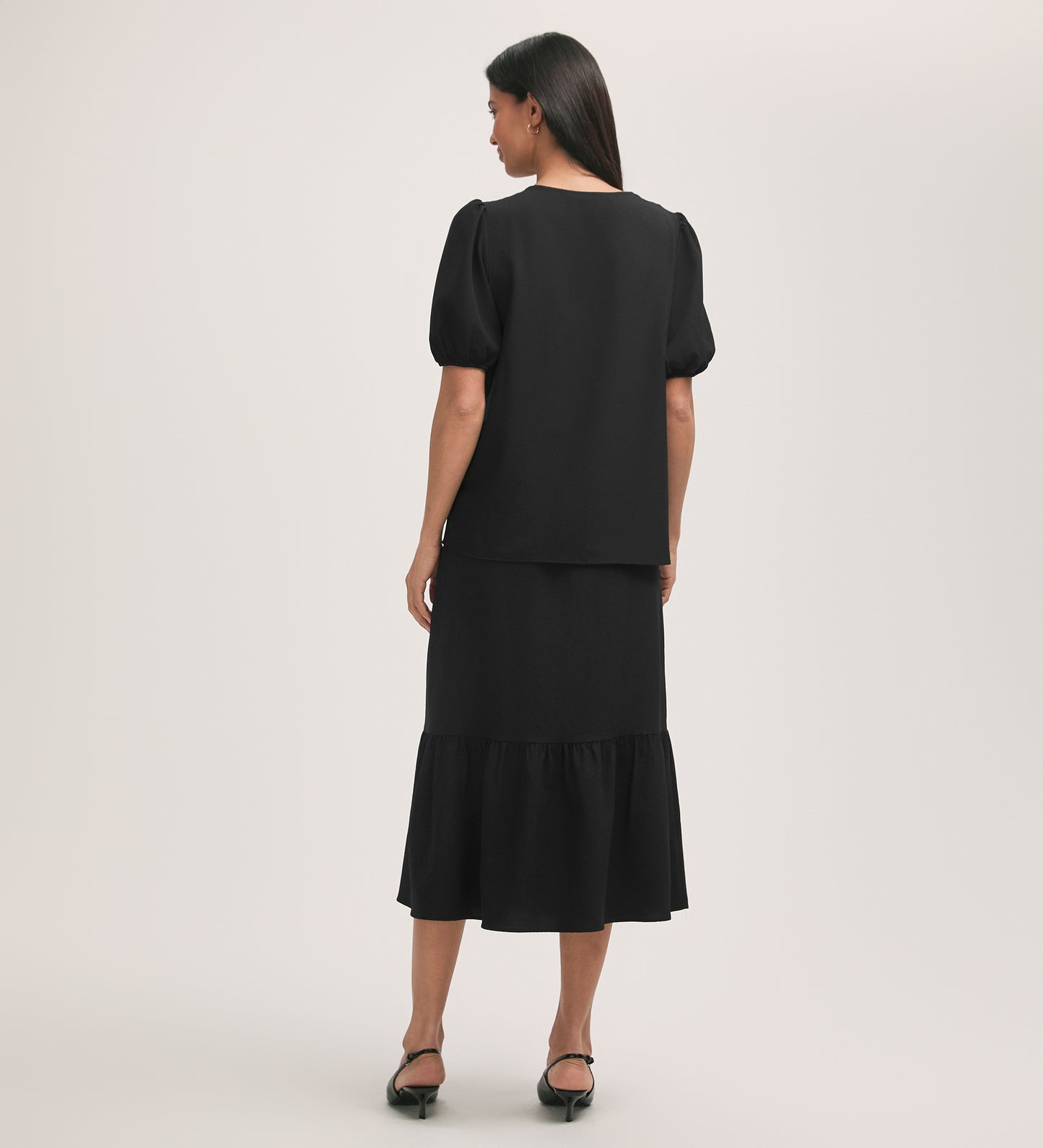 A woman with straight dark hair faces away, wearing the Fleur Black Crepe Short Sleeve Button Up Blouse and a black ruffled midi skirt. She completes the look with black heeled sandals against a plain light background.