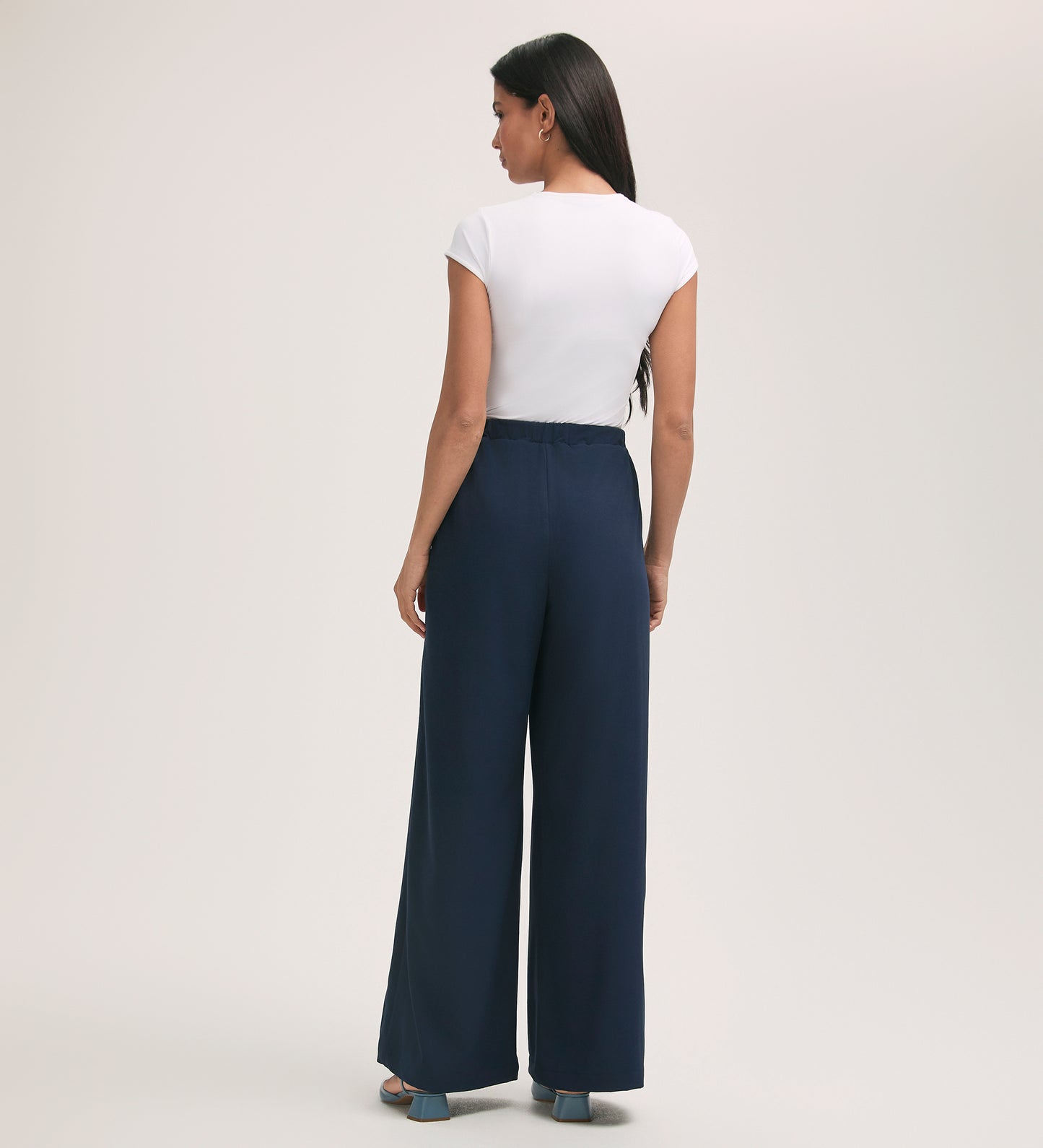 A woman stands facing away, wearing a white short-sleeve top and the Nat Navy Crepe Wide Leg Trousers with blue heeled sandals, set against a plain light background.