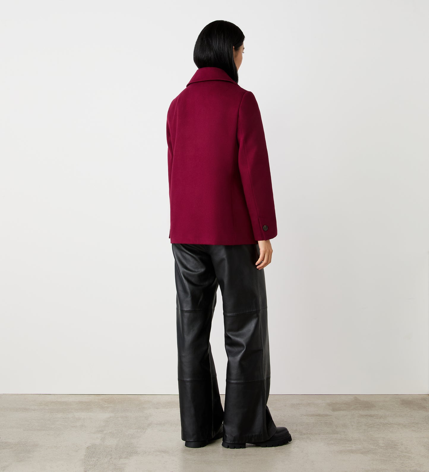 A person with short black hair stands facing away on a concrete floor against a white wall, wearing the Gemma Burgundy Double Breasted Jacket and loose black leather pants.