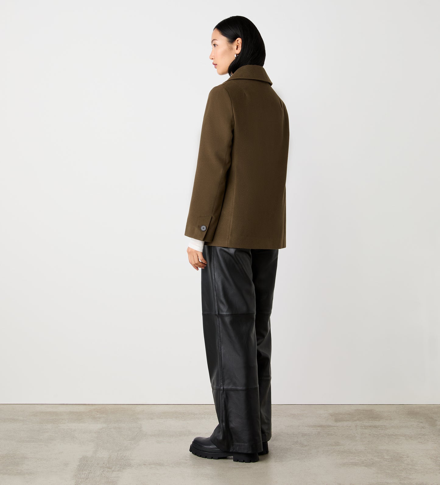 A woman with dark hair faces away from the camera in a minimalist setting, wearing the Gemma Khaki Double Breasted Jacket, black leather pants, and black boots against a light gray wall and concrete floor.