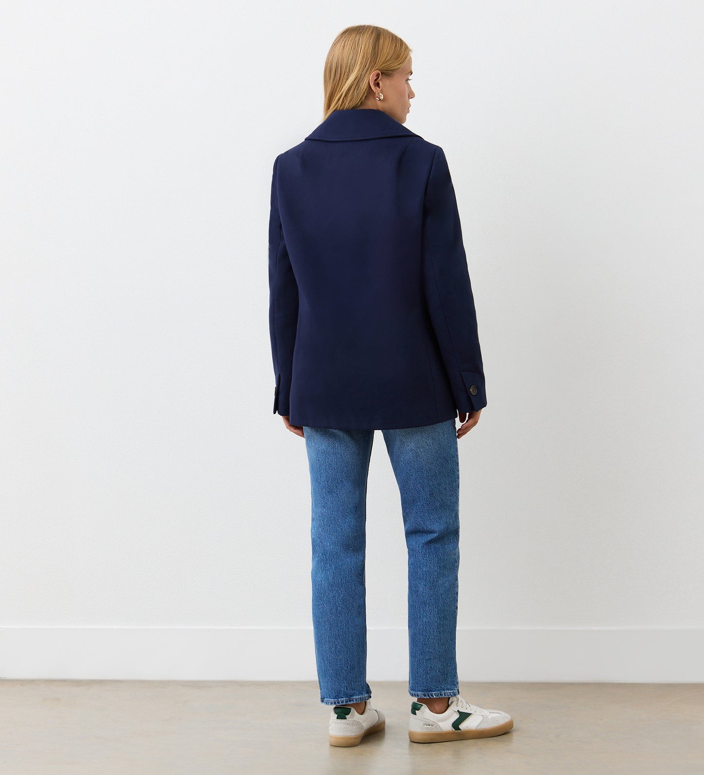 A person with shoulder-length blonde hair, wearing the Gemma Navy Double Breasted Jacket, blue jeans, and white sneakers, stands facing away from the camera in a minimal indoor space with a light floor and white wall.