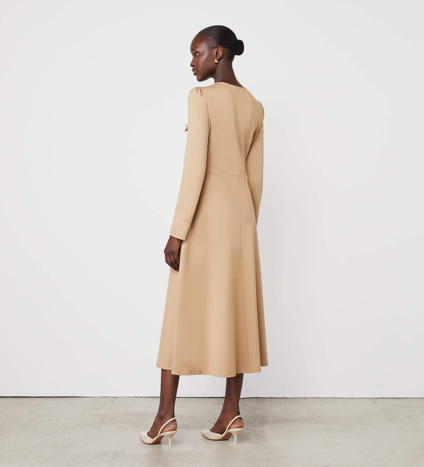 A woman stands against a white wall, wearing the Gilly Camel Ponte Jersey Midi Dress with long sleeves and gold button detail, paired with beige heeled shoes. She faces away but turns slightly to the side, her hair styled in a neat bun.