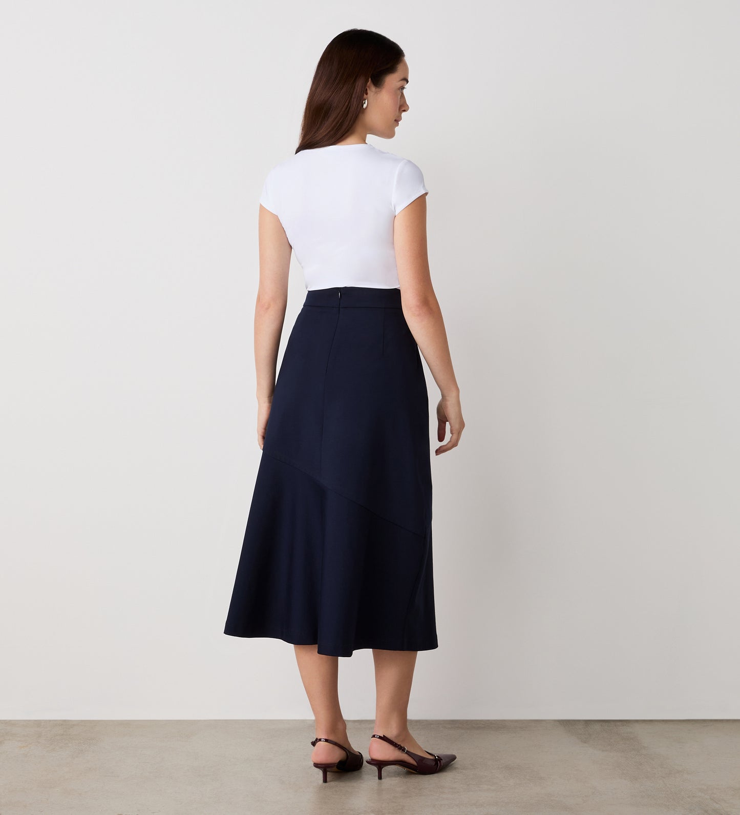 A woman with long brown hair stands indoors facing away, wearing the Grace Navy Ponte Jersey Midi Skirt and a white short-sleeve top, on a light floor against a plain white wall. The skirt features an elasticated waistband.