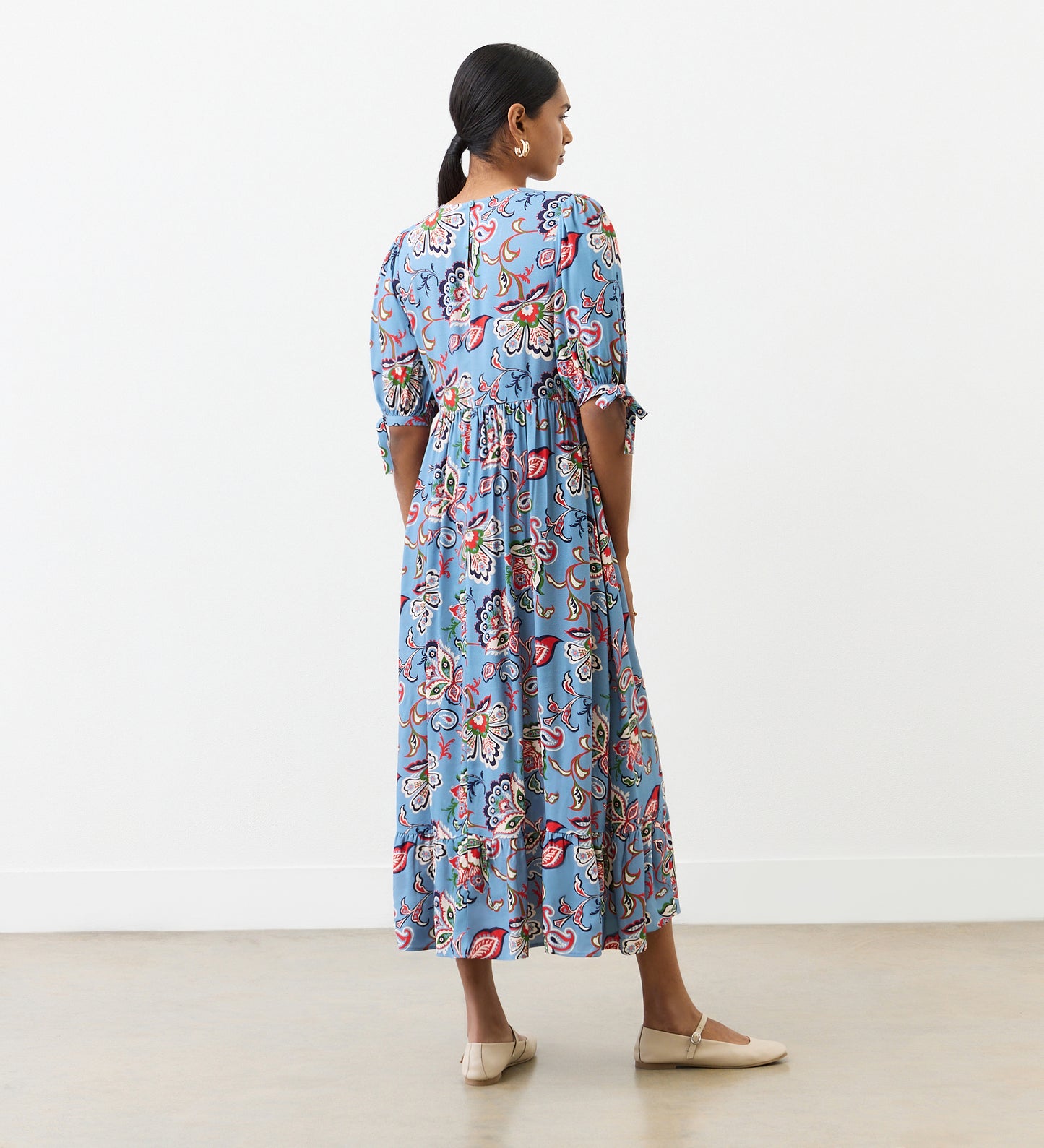 A woman with dark hair tied back stands indoors facing away, wearing the Gwen Blue Floral Midi Smock Dress with elbow-length sleeves and a colorful floral pattern, paired with beige flats. She stands on a plain floor against a white wall.