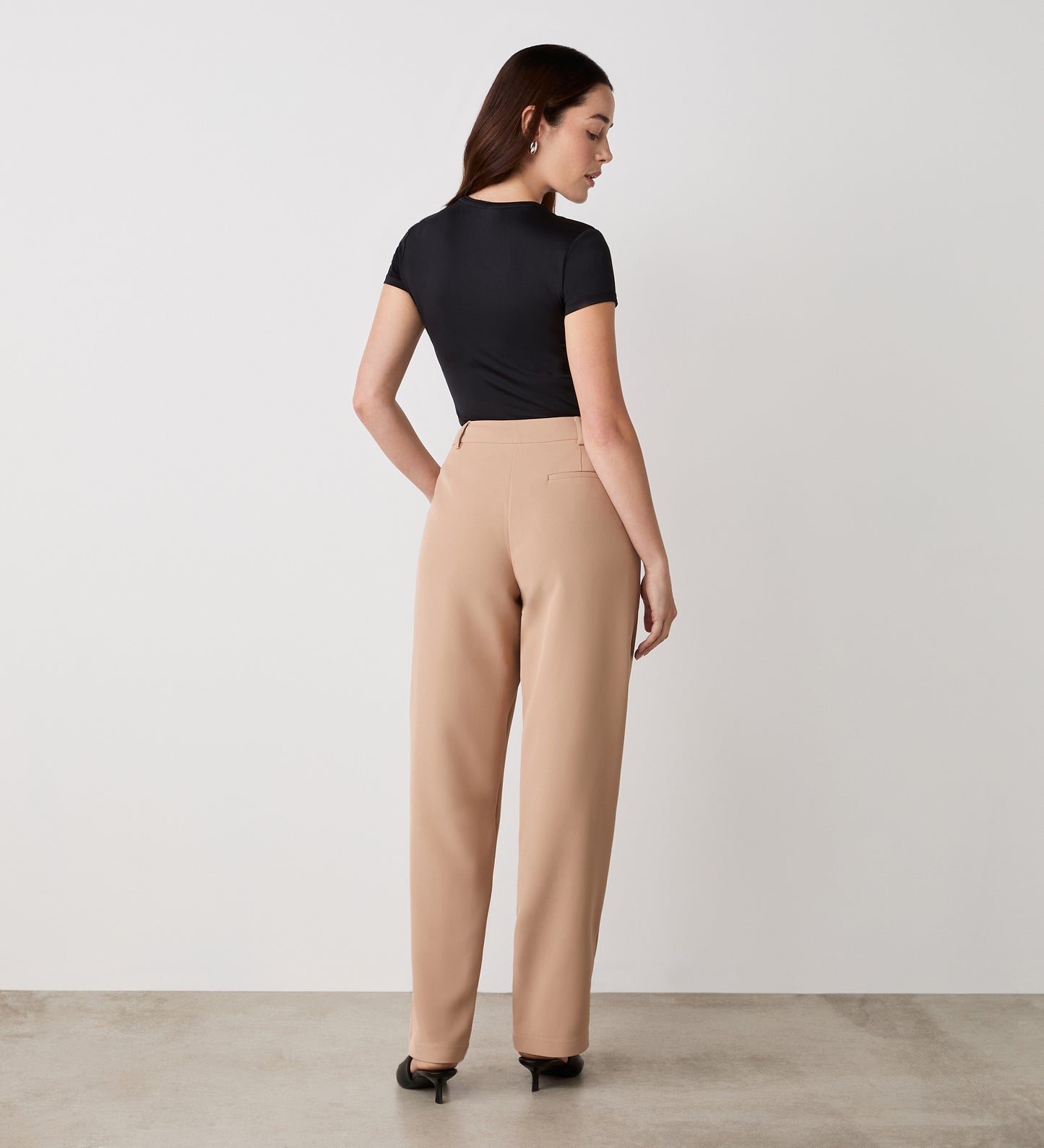 A woman stands facing away, wearing a fitted black top and Harper Camel Straight Leg Trousers—wardrobe staples—paired with black heels against a plain white wall and concrete floor.