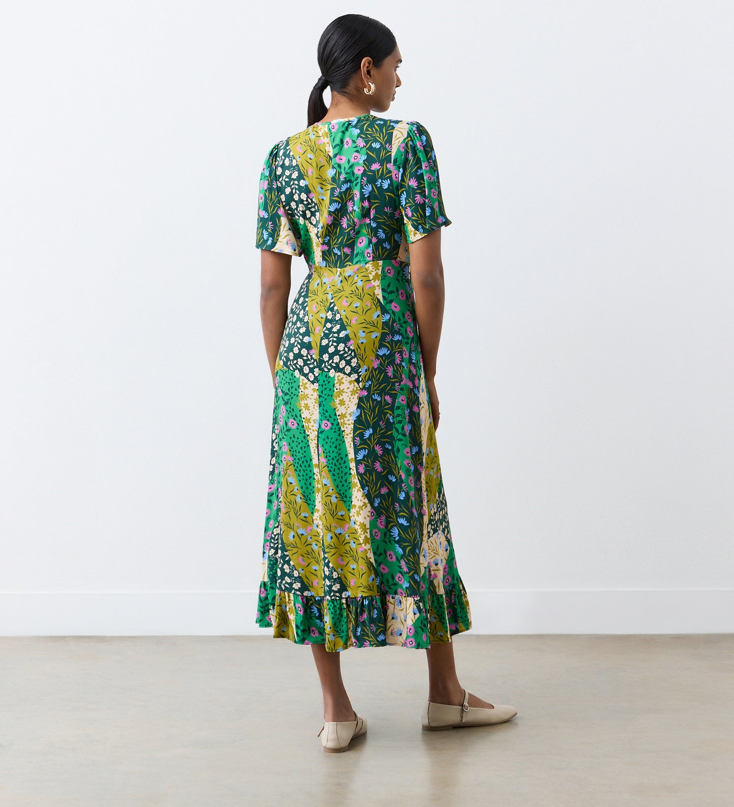 A woman stands indoors facing away, wearing the Isabella Green Patchwork Floral Tie Neck Crepe Midi Dress with an empire waist and ruffled hem. Her hair is tied back, paired with light-colored flats against a plain background.