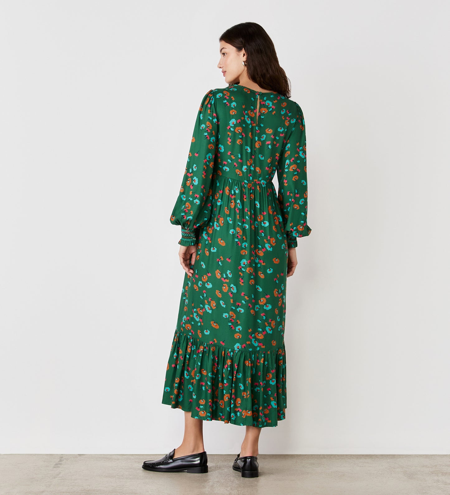 A woman, seen from behind, wears the Isha Green Floral Midi Dress with long sleeves, a shirred bodice, and tiered hem, styled with black loafers against a white background.