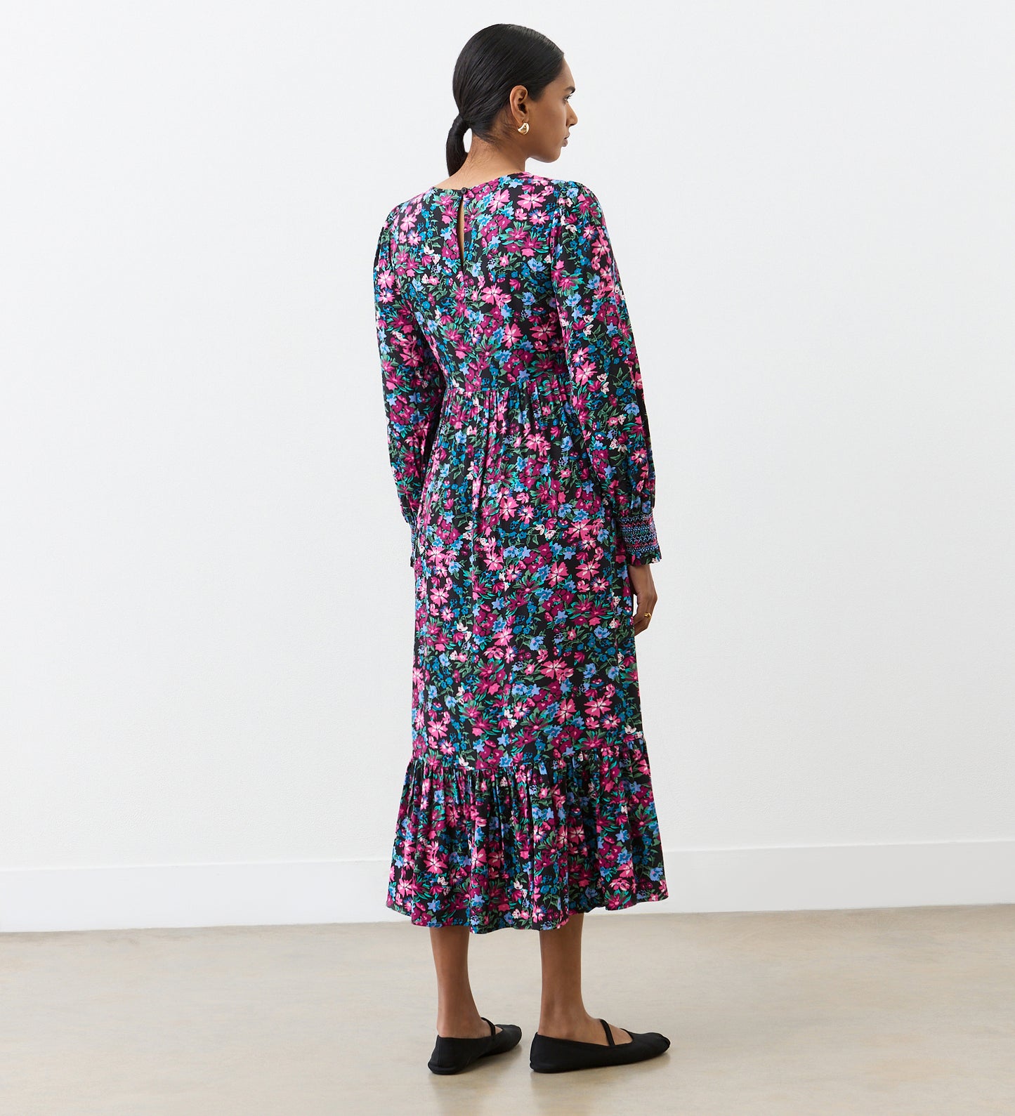 A woman with dark hair pulled back stands indoors facing away, wearing the Isha Pink Floral Midi Dress with a tiered hem and shirred bodice, paired with black flat shoes against a plain white background.