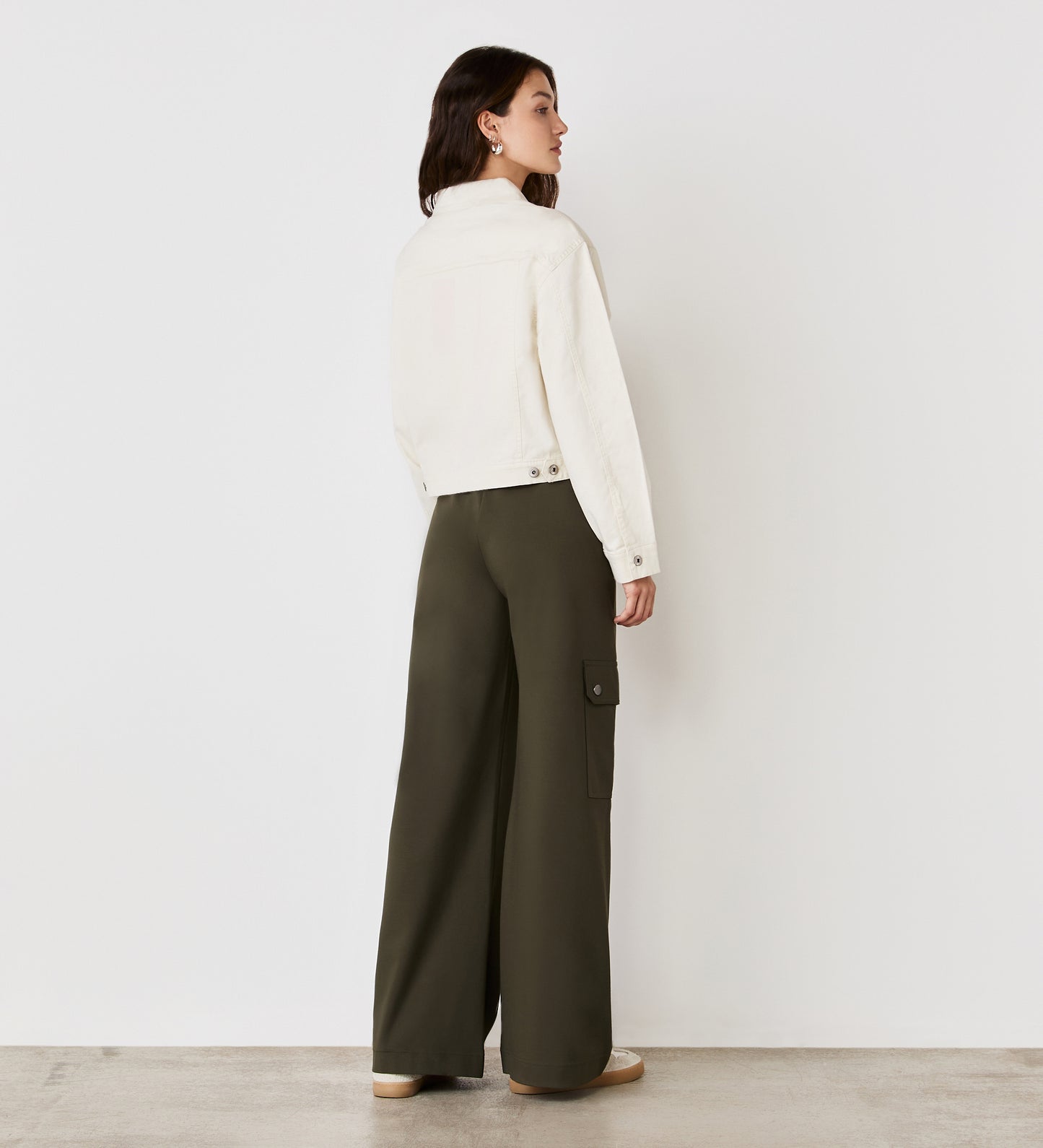 A woman faces away, wearing the Isobel Ivory Denim Jacket with a flattering fit, paired with olive green wide-leg cargo pants and beige shoes, against a white wall and concrete floor.