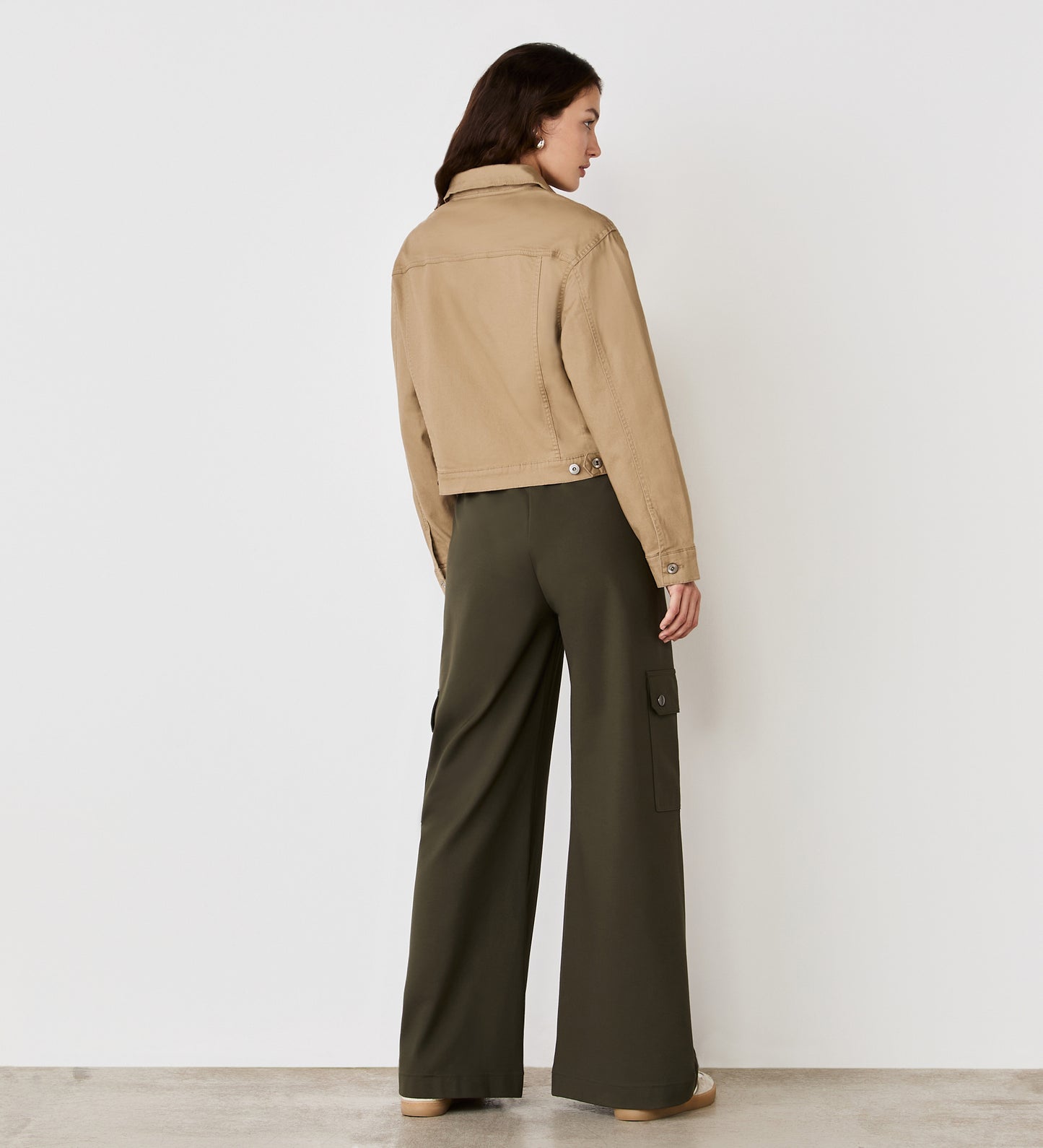 A woman stands facing away from the camera, wearing the Isobel Mocha Denim Jacket and olive green wide-leg pants with side pockets, against a plain white wall background.