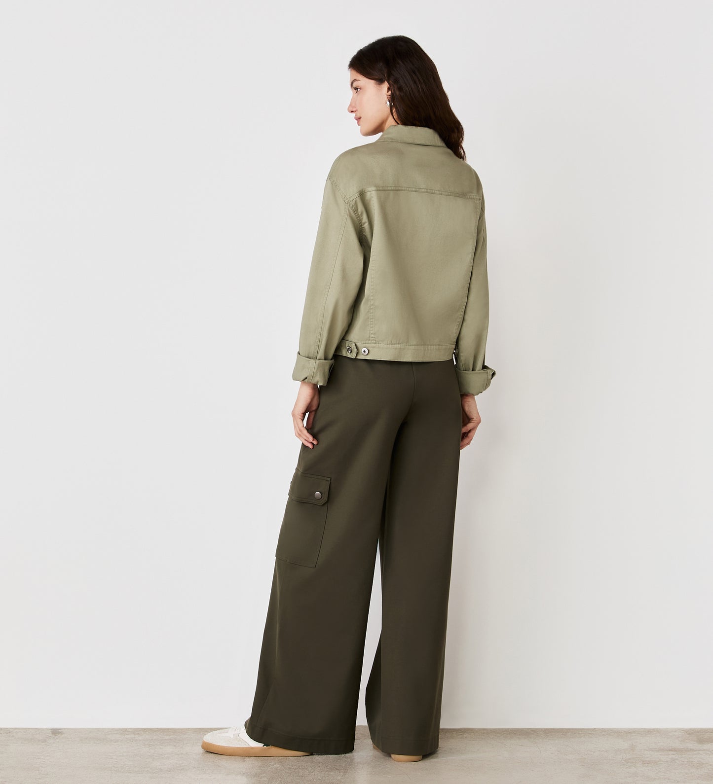 A woman faces away from the camera, modeling the Isobel Sage Denim Jacket with a flattering fit and wide-leg dark green cargo pants with side pockets, set against a plain white background.