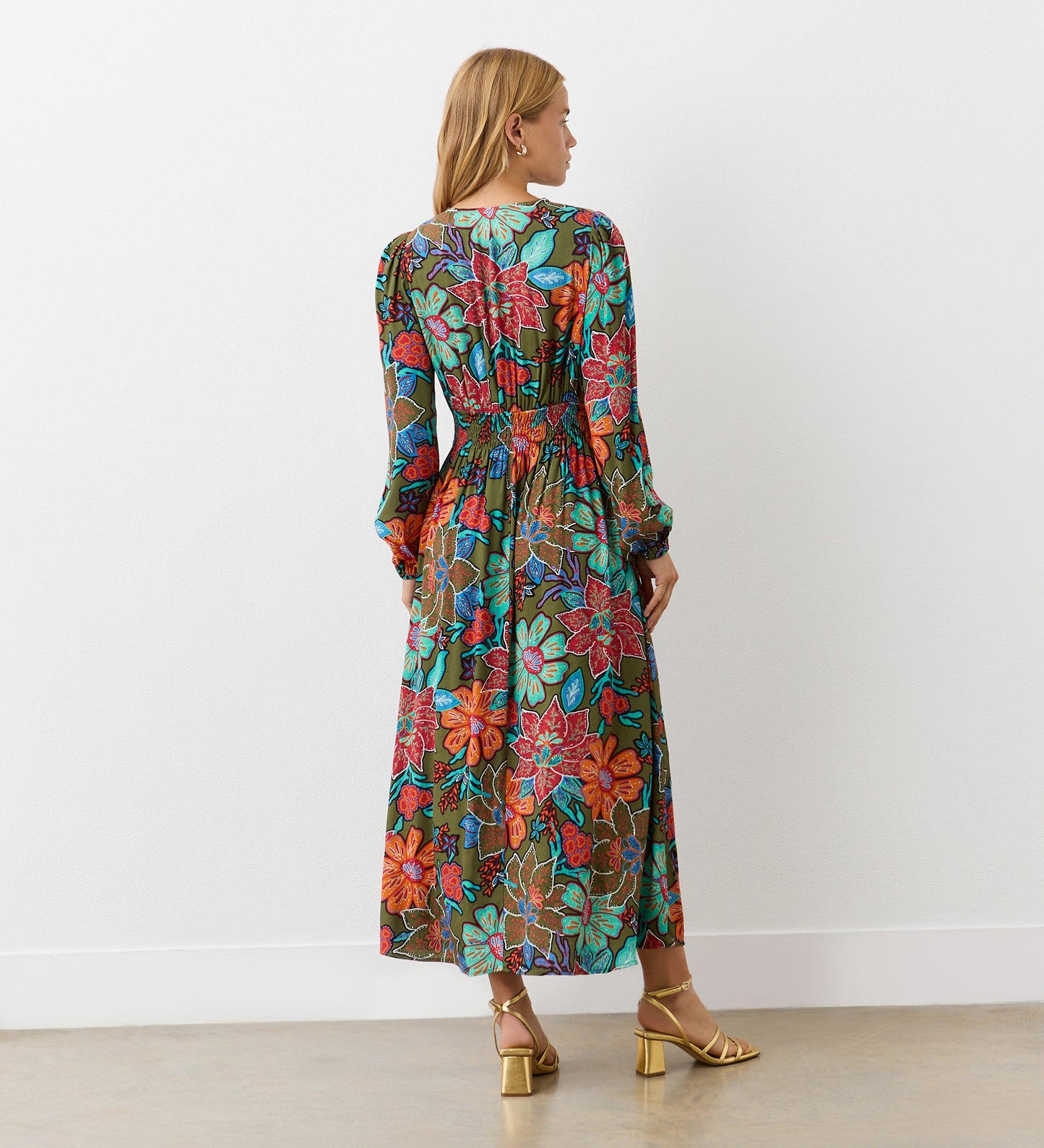 A woman with blonde hair stands facing away in the Janis Khaki Floral Crepe Midi Dress with full-length sleeves, paired with gold heeled sandals. The background features a plain white wall and a light-colored floor.