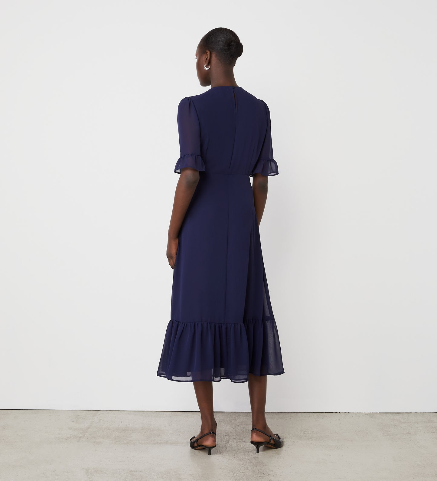 A woman stands with her back to the camera, wearing the Jennifer Navy Chiffon Midi Dress featuring an empire waist, short ruffled sleeves, and a ruffled hem. She pairs it with black sandals; her hair is in a bun against a white wall.