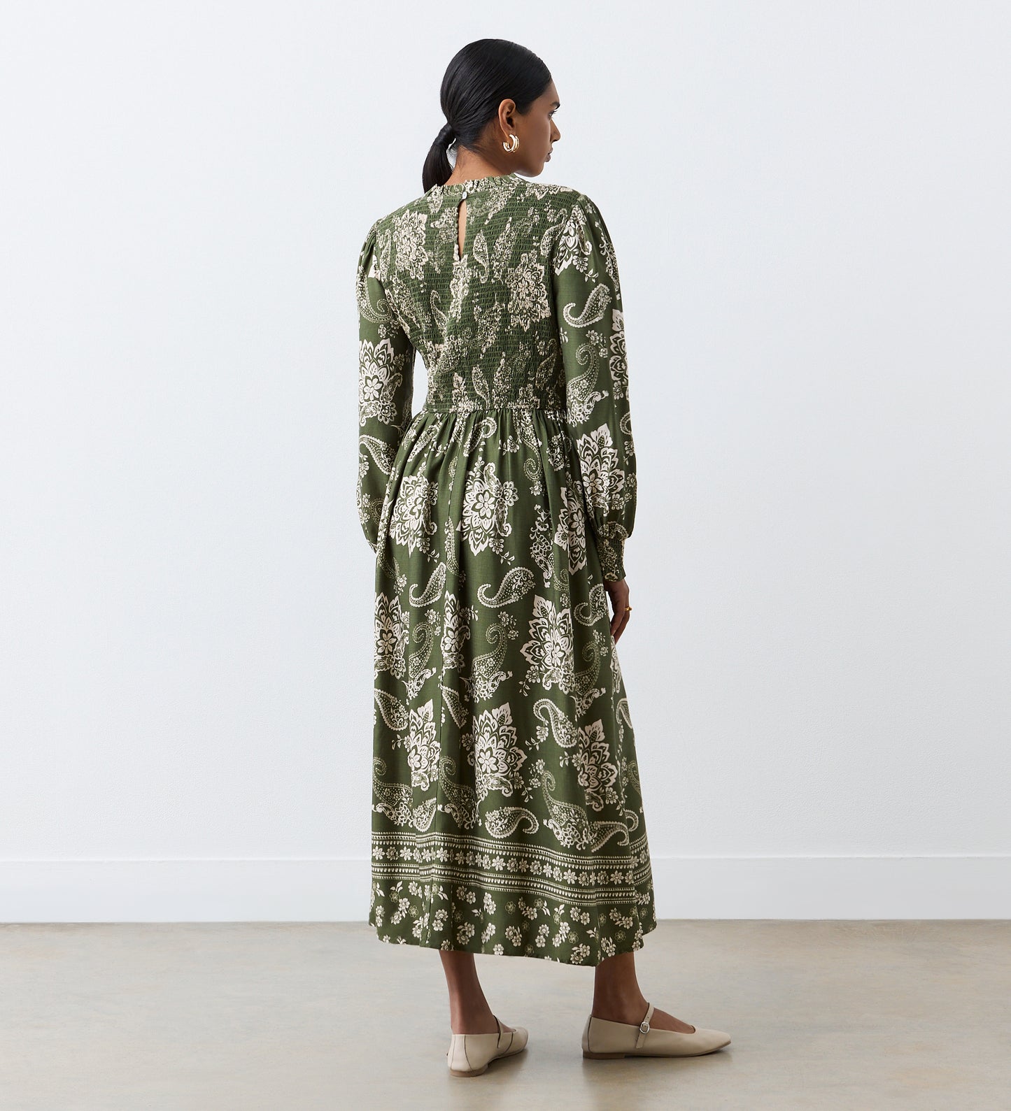 A woman stands indoors facing away, wearing the Jovie Green Paisley Midi Dress with elegant sleeves and paisley print. Her hair is tied back, and she completes the look with beige flats against a plain white background.
