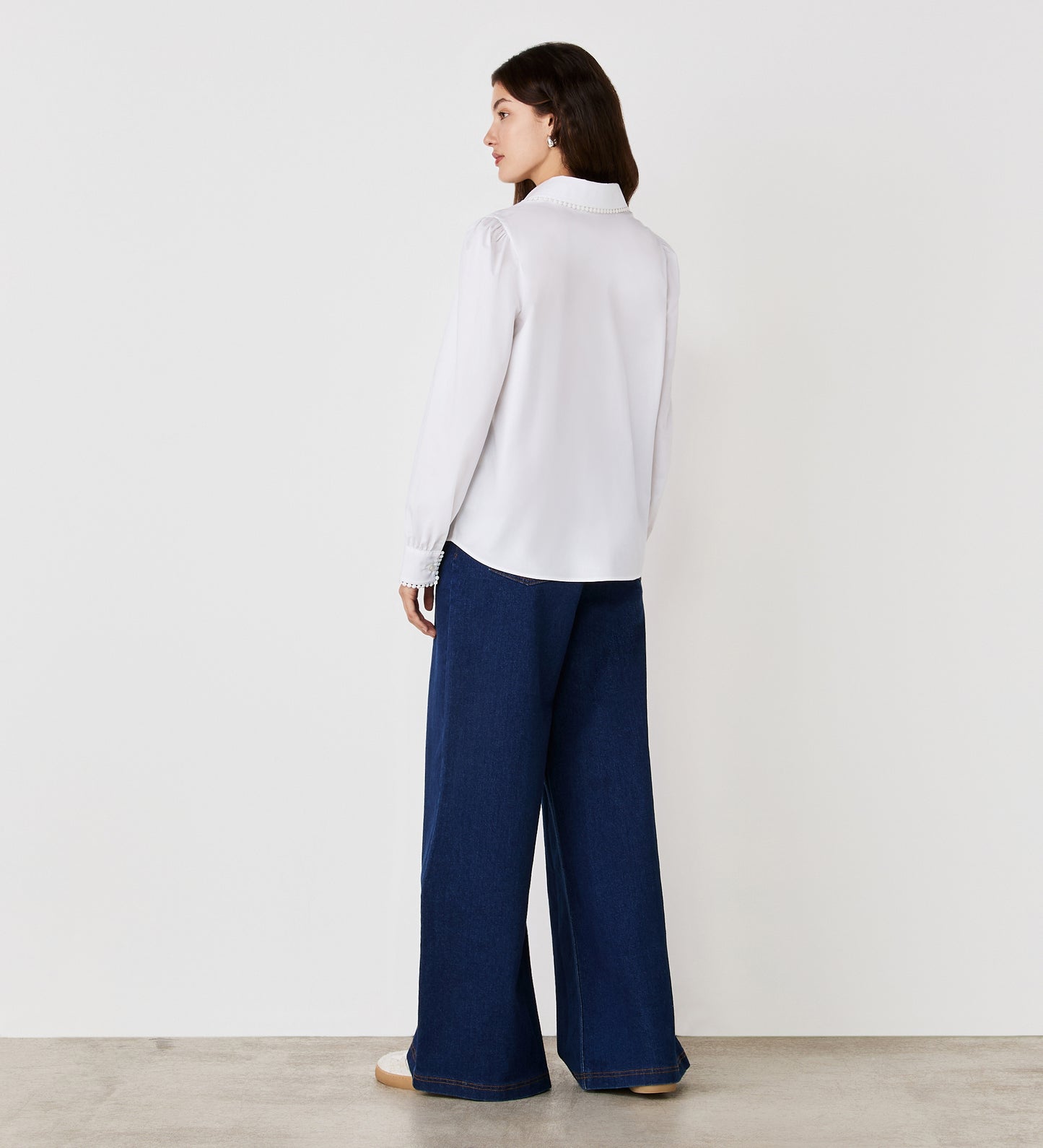 A woman stands with her back to the camera, wearing the Judith White Cotton Rich Shirt and wide-leg blue jeans. She is on a light-colored floor in front of a plain white wall.