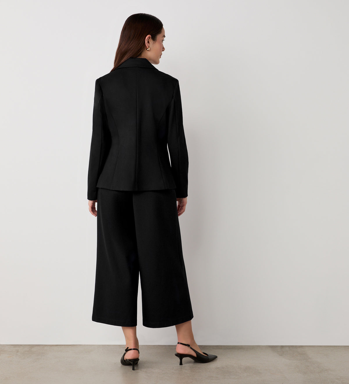 A woman faces a white wall in a minimalist setting, wearing the Jules Black Ponte Jersey Blazer with princess seams, paired with wide-leg black pants and black heels; only her back is visible.