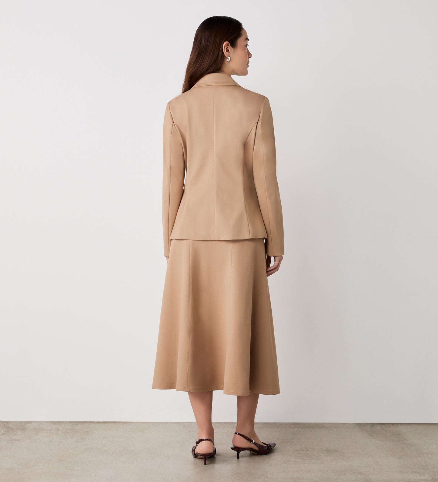 A woman with long brown hair stands facing away, wearing the Jules Camel Ponte Jersey Blazer and matching midi skirt with heeled sandals, set against a plain white wall and concrete floor.
