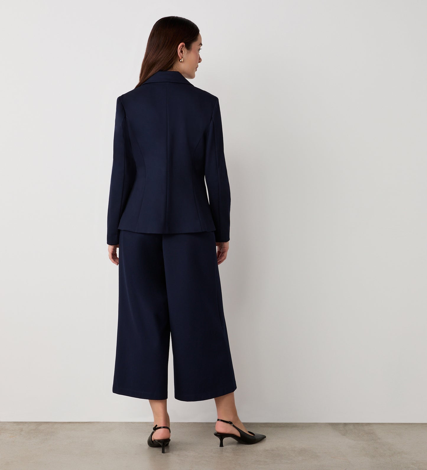 A woman faces away from the camera, wearing the Jules Navy Ponte Jersey Blazer with matching wide-leg cropped trousers and black heels, against a plain white wall and light floor.
