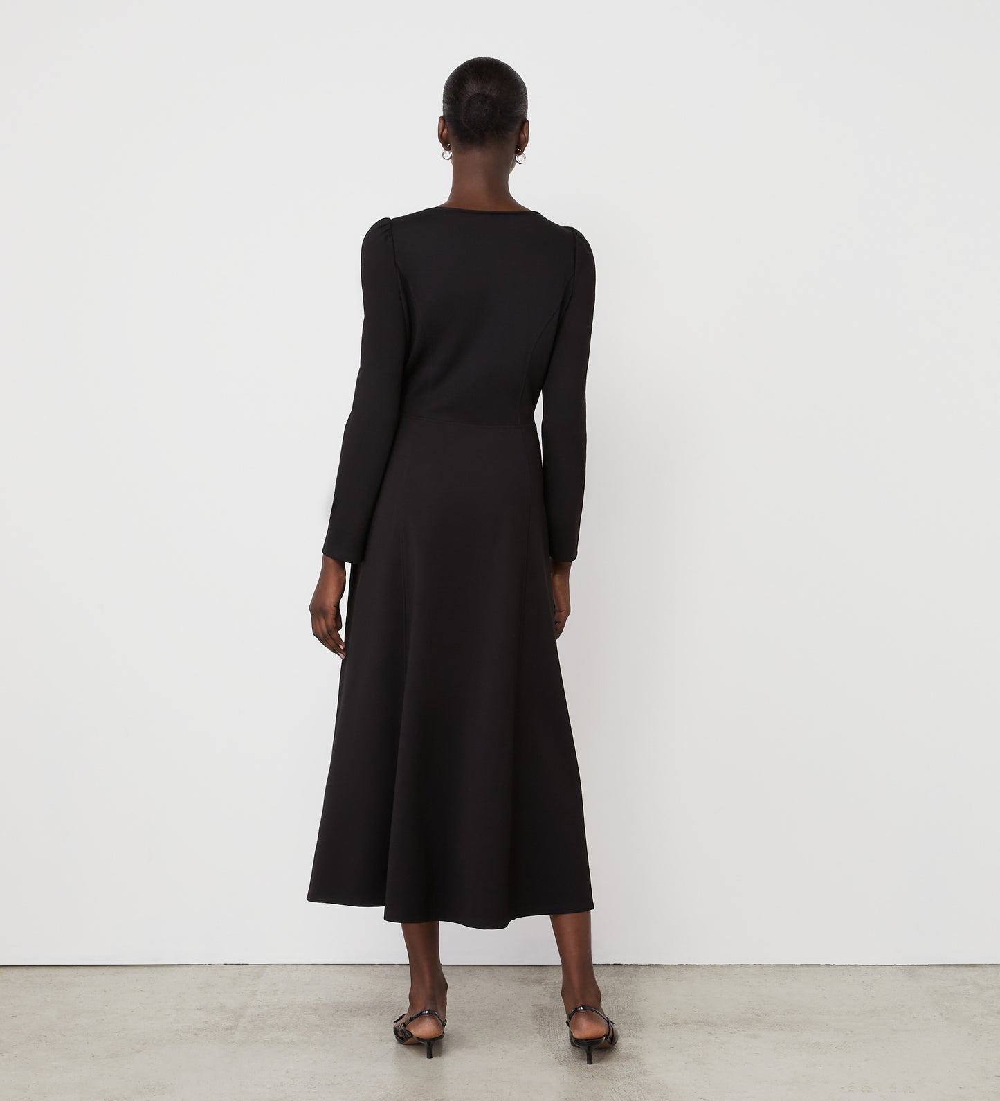 A woman faces away from the camera, wearing the Kelly Black Ponte Jersey Midi Dress with long sleeves and subtle seams, paired with black heeled sandals against a plain white wall and concrete floor.
