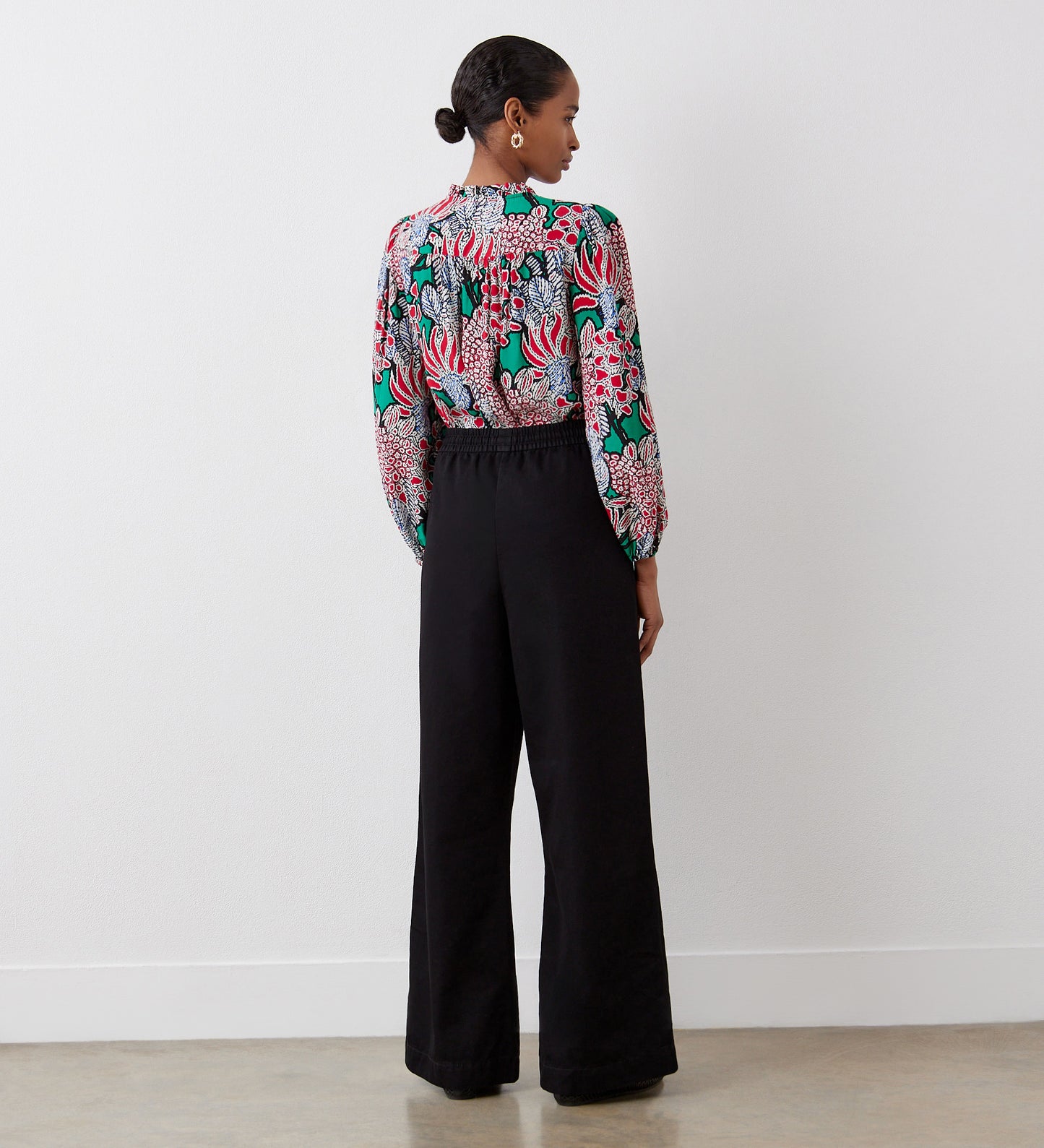 A woman with short, dark hair stands against a white wall wearing high-waisted Dixie Black Denim Wide Leg Trousers and a colorful floral long-sleeve blouse, facing away.