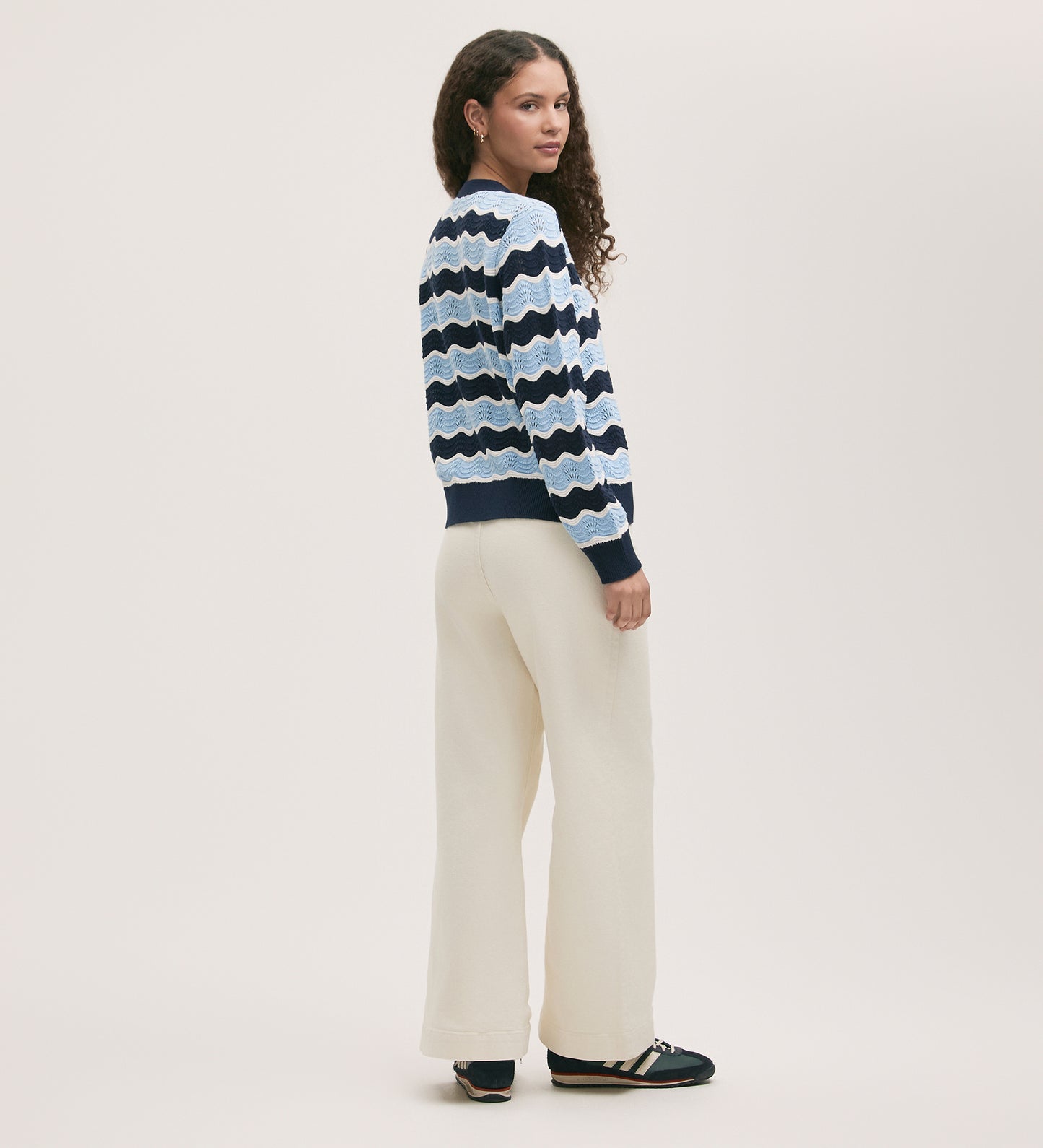 A woman with long curly hair stands sideways, looking back at the camera. She wears the Kate Blue Stripe Crochet Cardigan, cream wide-leg pants, and black sneakers with white stripes.