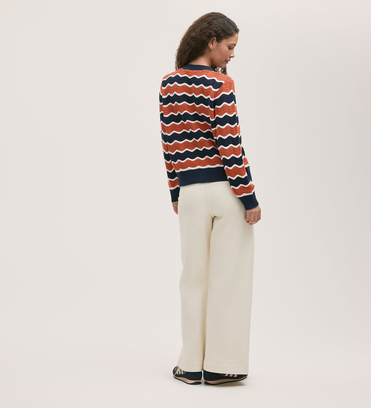 A woman faces away, wearing the Kate Orange Stripe Crochet Cardigan—featuring relaxed orange stripes—paired with cream wide-leg pants and dark sneakers against a plain light background.