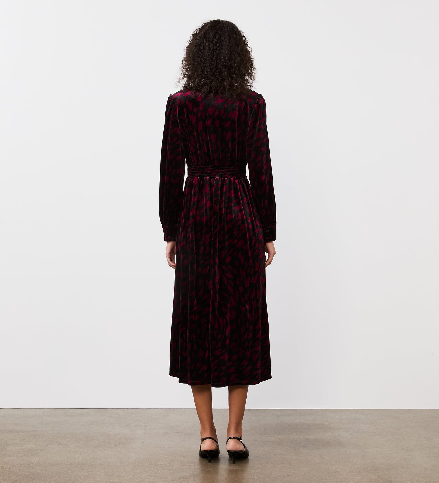 A woman with curly hair faces away, modeling the Layla Burgundy Animal Velvet Midi Dress—its long sleeves and rich red animal pattern drape elegantly as she poses on a bare concrete floor against a plain white wall.