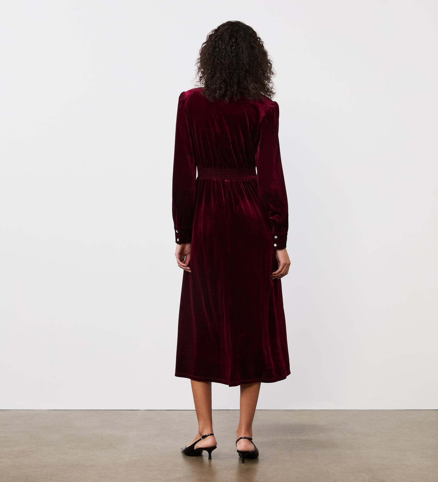 A woman with curly hair, seen from behind, wears the Layla Burgundy Velvet Midi Dress with black kitten-heel shoes, standing on a concrete floor against a plain white wall.