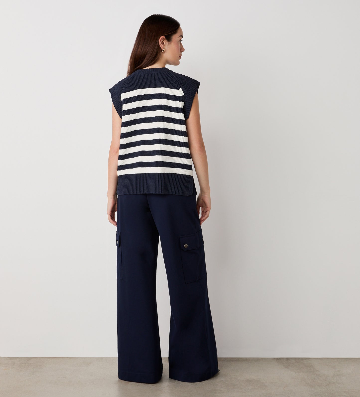 A woman stands facing away, wearing the Lexi Ivory Striped Sweater Vest and navy wide-leg cargo pants. The heavy knit adds texture, while her straight brown hair contrasts with the white wall and concrete floor.