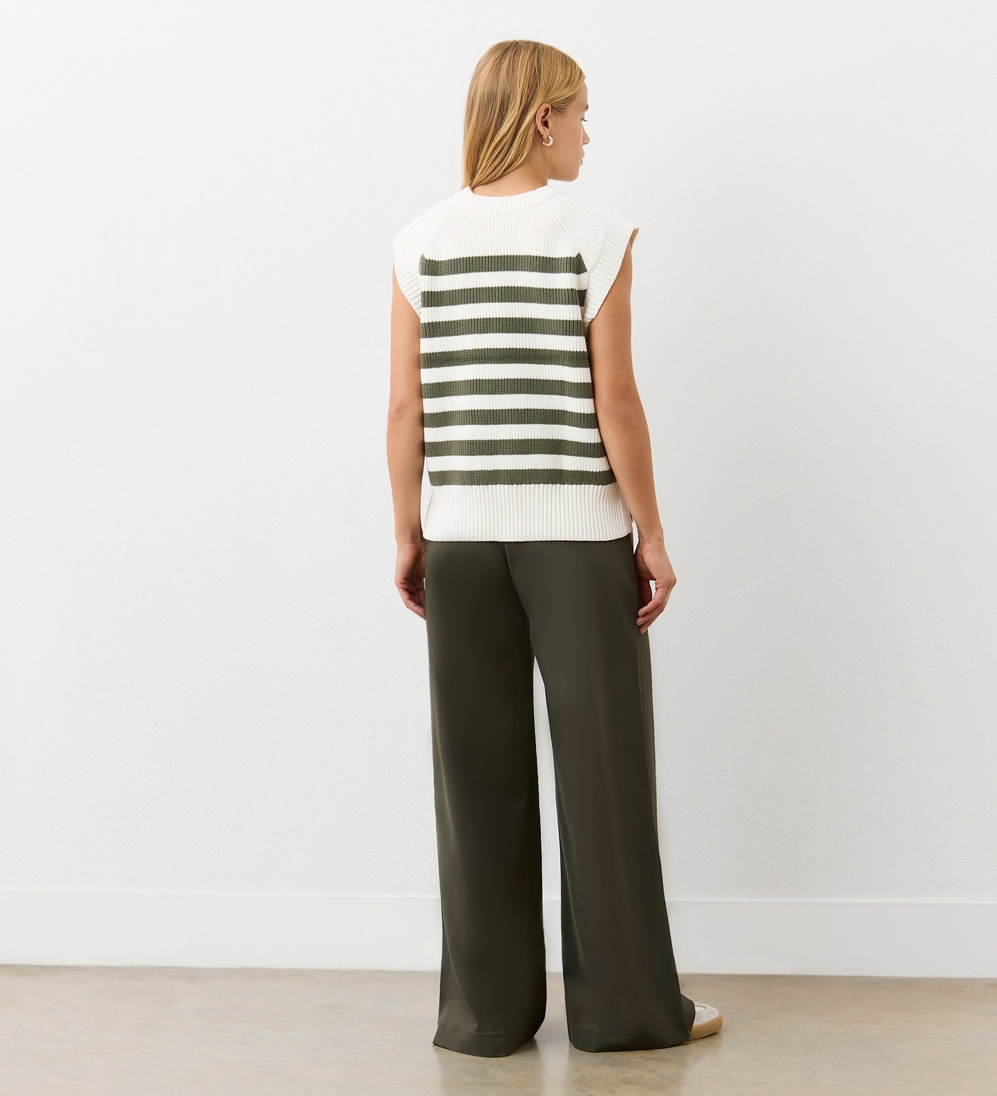 A woman with straight blonde hair wears the Lexi Khaki Striped Sweater Vest, featuring wide dark green stripes on the back, paired with loose dark green pants as she faces a white wall.